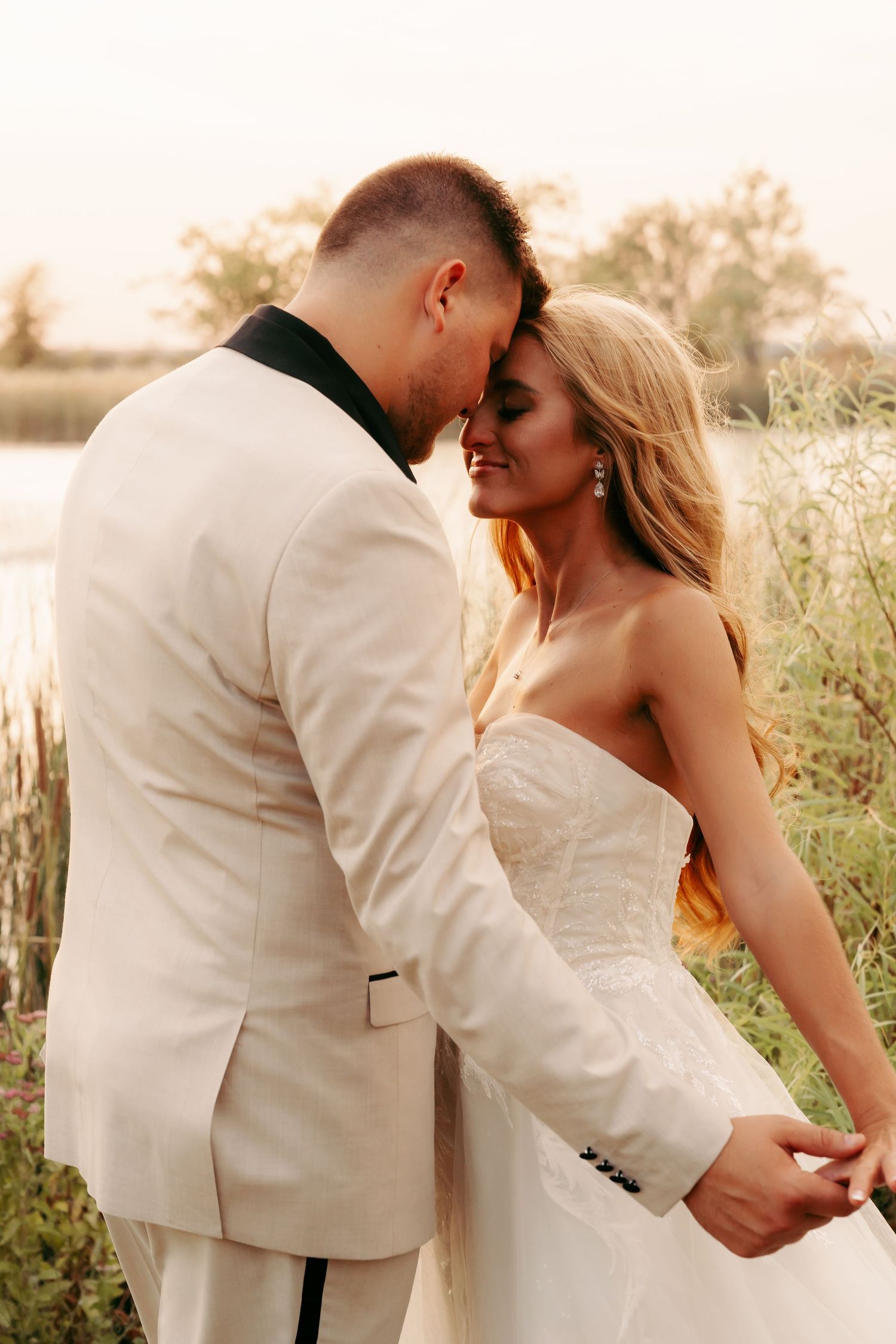 A romantic sunset wedding portrait of a couple in white attire embracing in a field of tall grass.