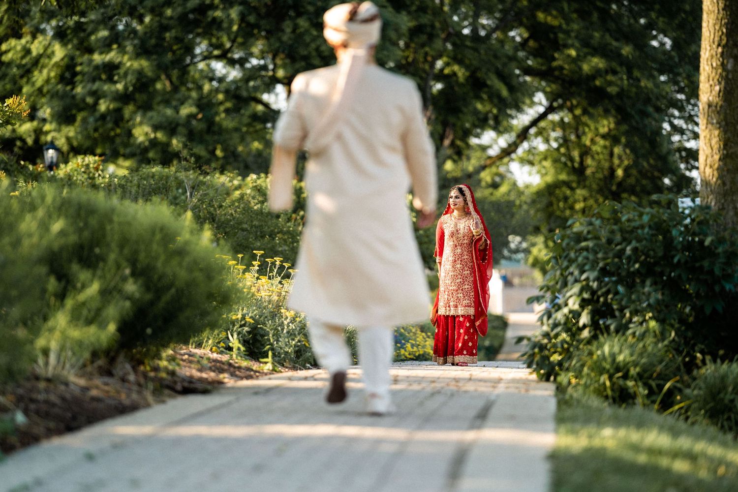 Indian groom's silhouette advances towards his bride in a resplendent red dress, amidst lush greenery and a paved path