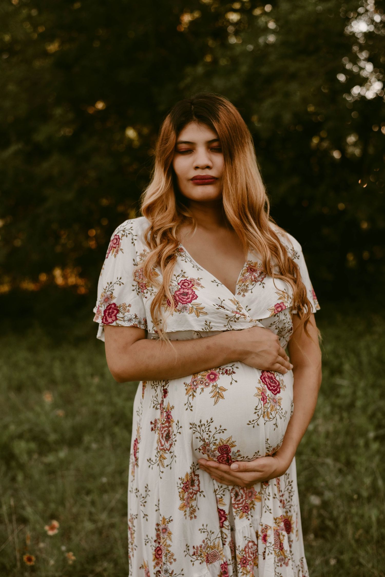 Maternity portrait in white floral dress with warm sunset lighting in natural outdoor setting.
