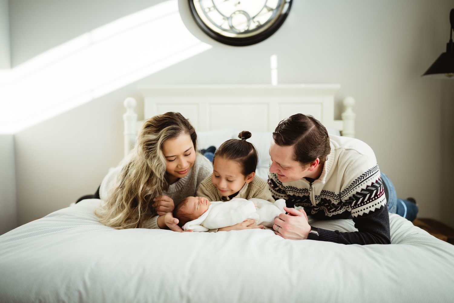 Family relaxes together on white bedding while wearing cozy winter sweaters.