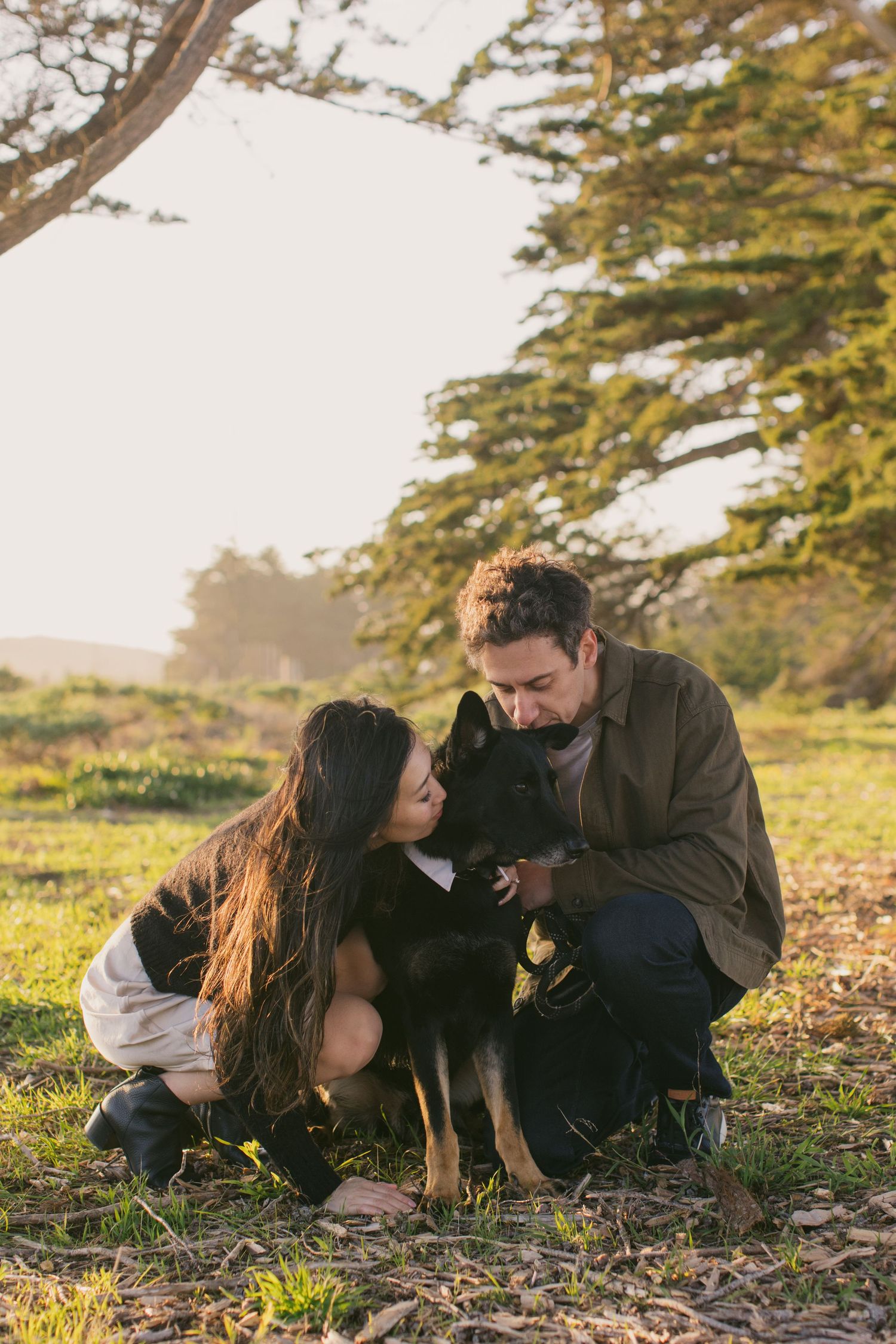 A couple sits with their black dog under large trees in a park setting.