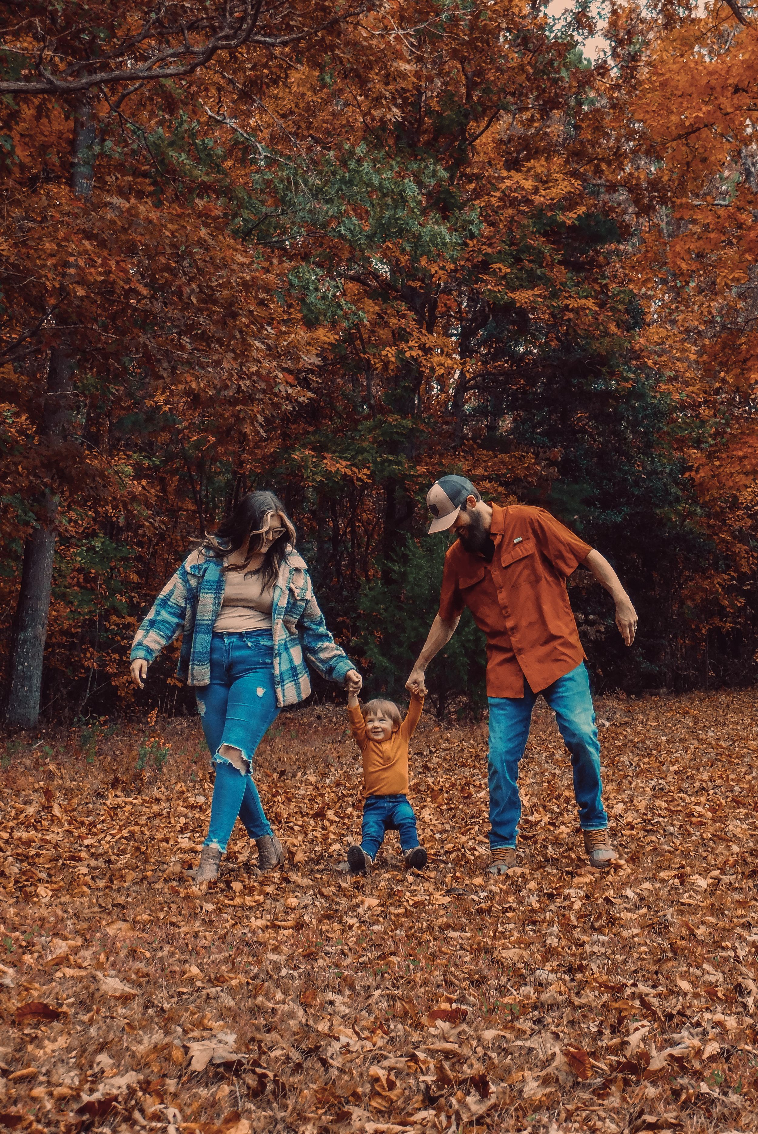 Three people playing in fallen autumn leaves surrounded by orange and red foliage in a forest setting.