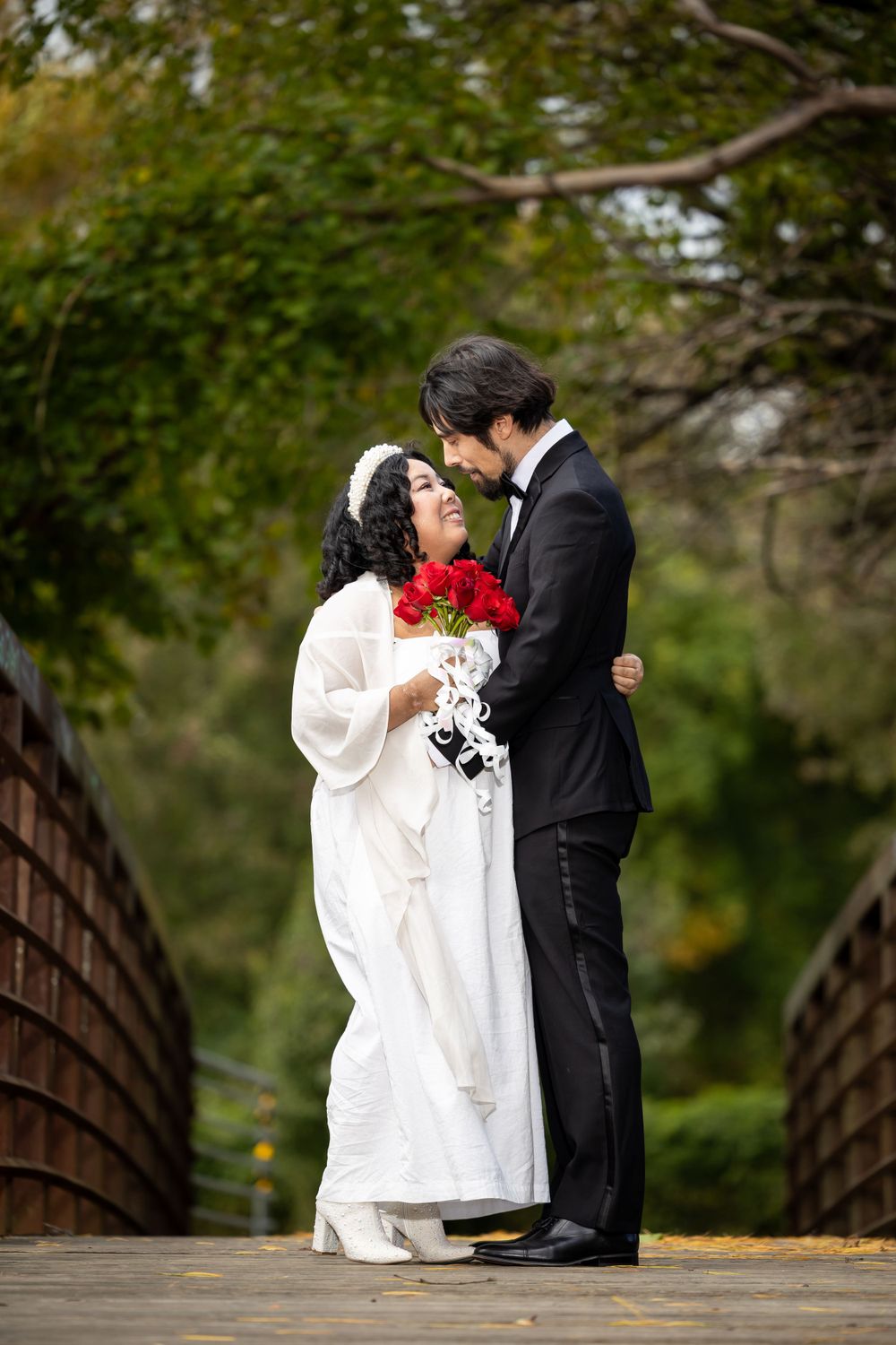 A tender moment between newlyweds on a wooden bridge with the bride holding red roses.