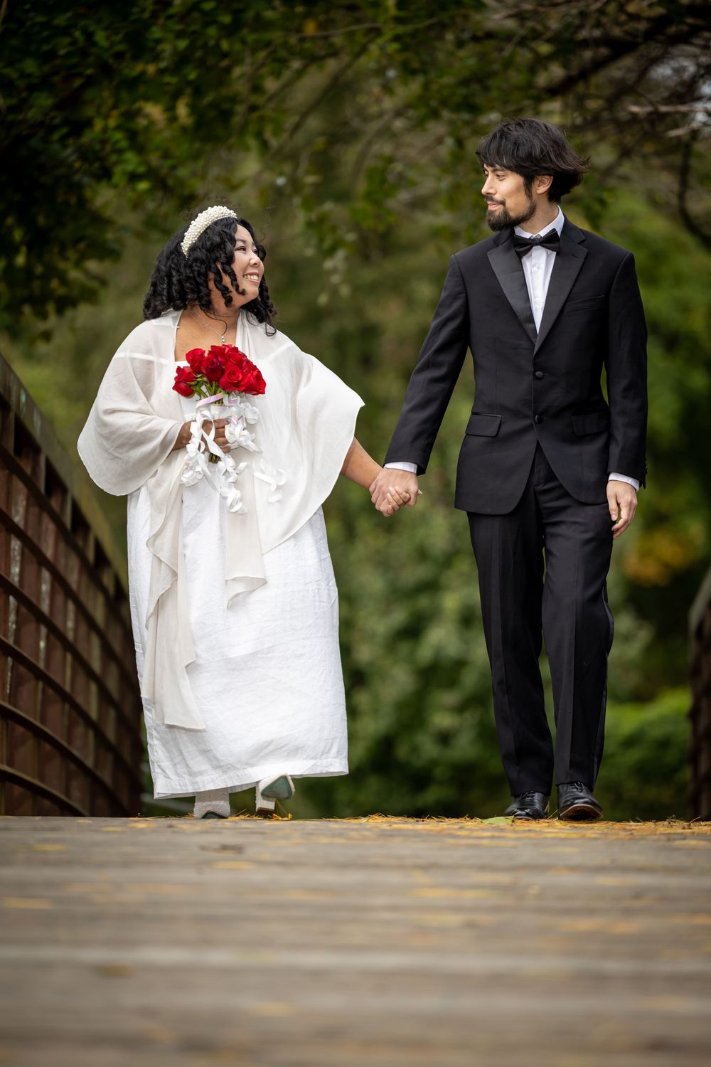 A couple in wedding attire walks hand in hand on a wooden bridge.