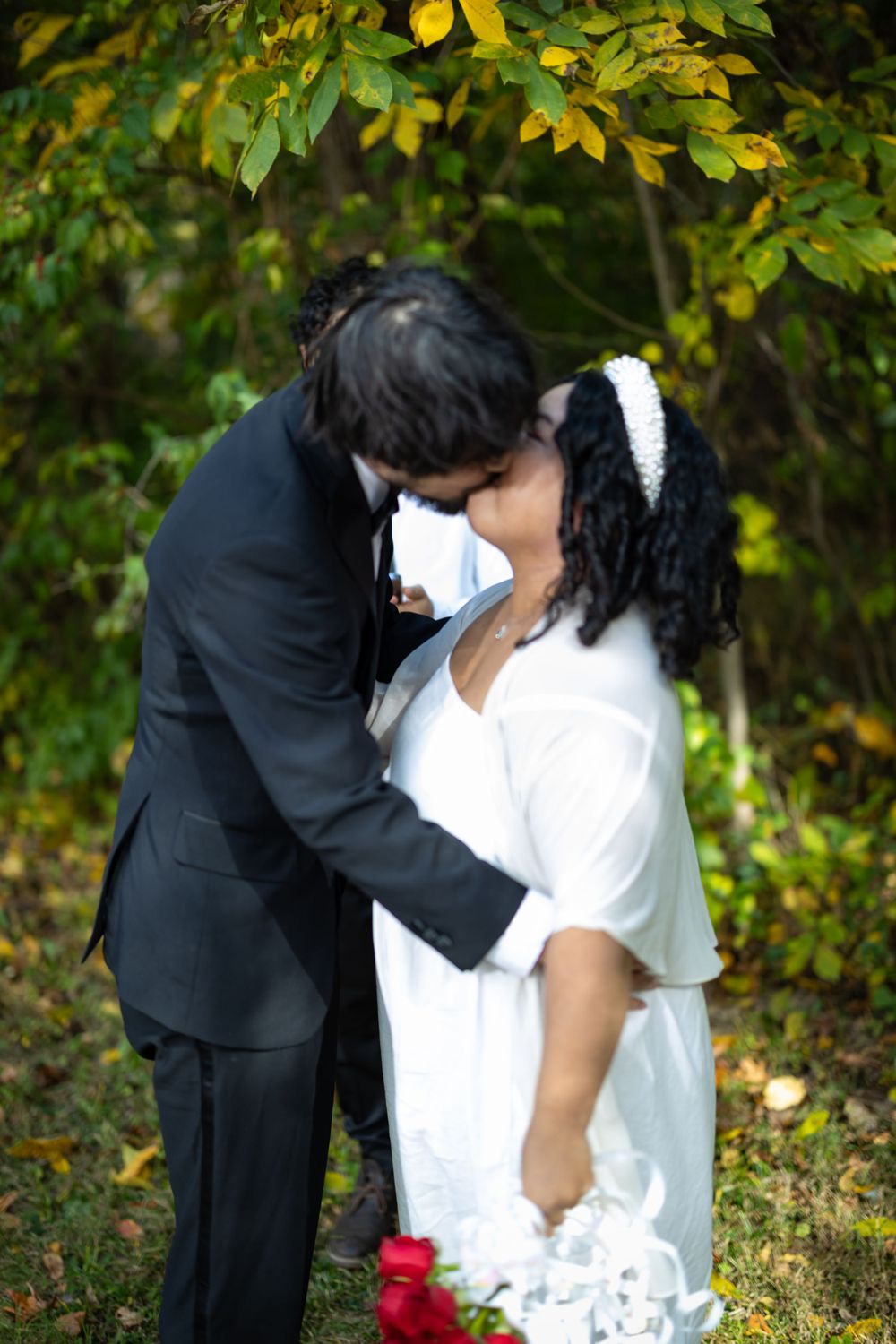 A romantic wedding couple embraces among fall foliage with the bride in white holding red roses.