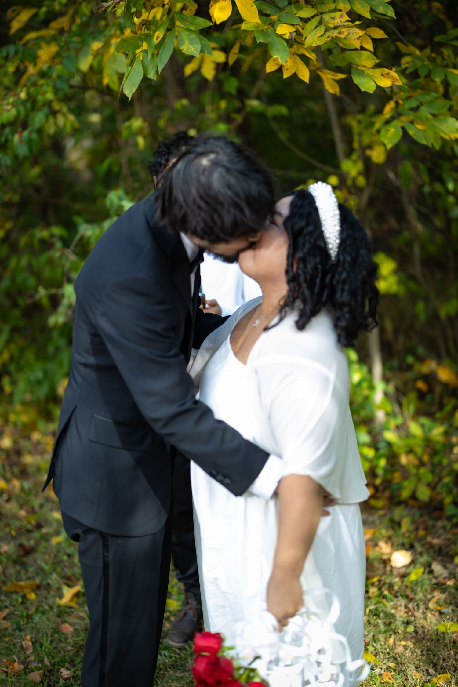 A romantic wedding couple embraces among fall foliage with the bride in white holding red roses.