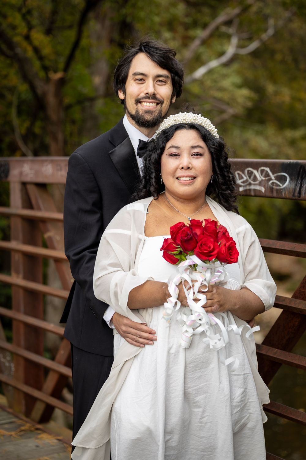 A wedding couple poses together on a wooden bridge with red roses and autumn scenery.