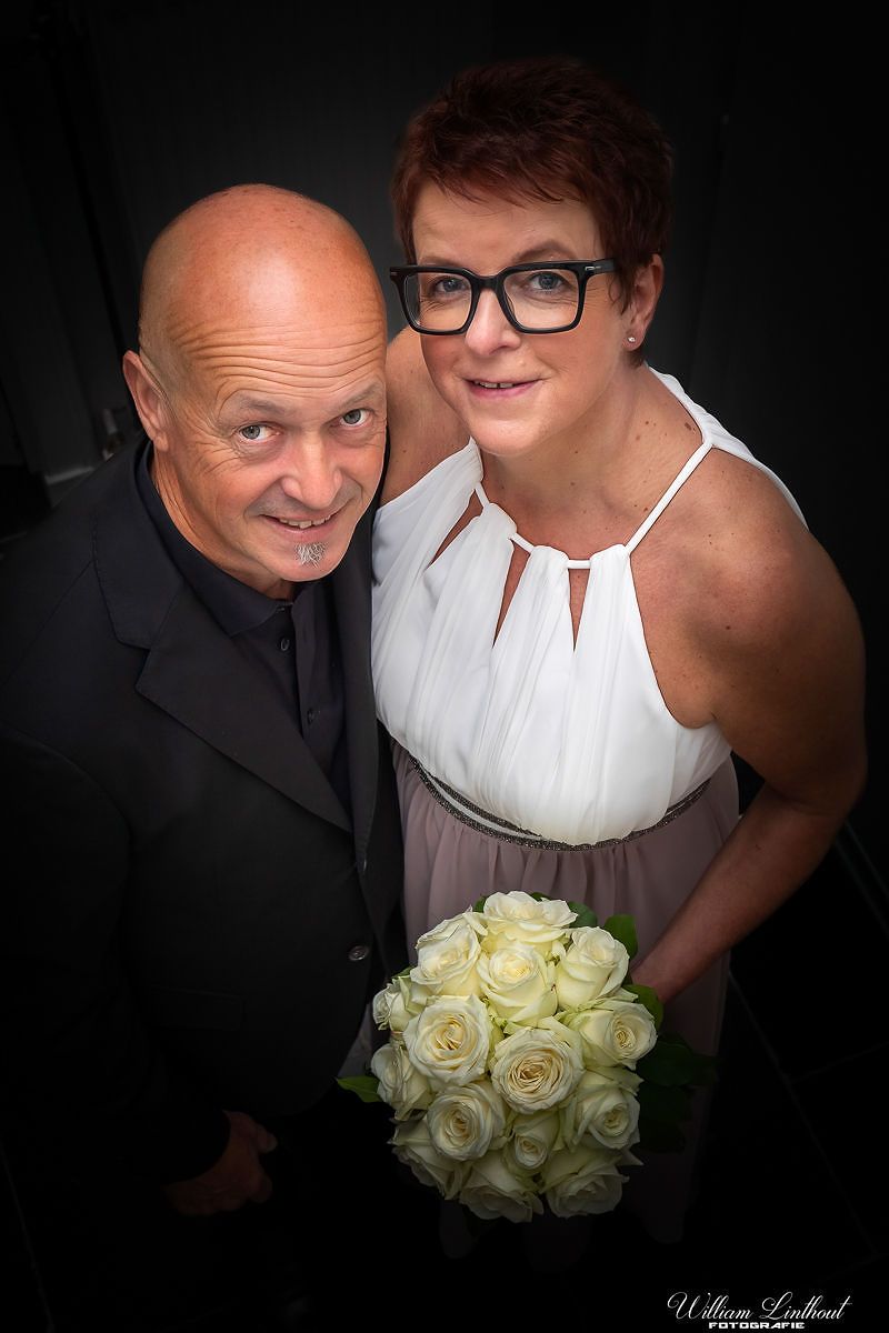 Smiling couple in formal attire pose together with white rose bouquet against dark background.