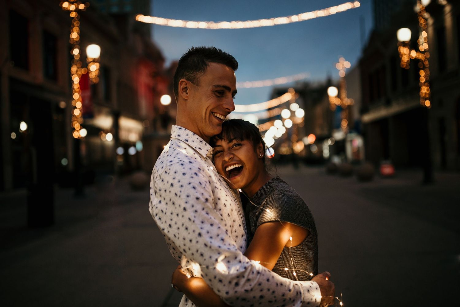 Couple hugs on a dark street with twinkle lights behind them