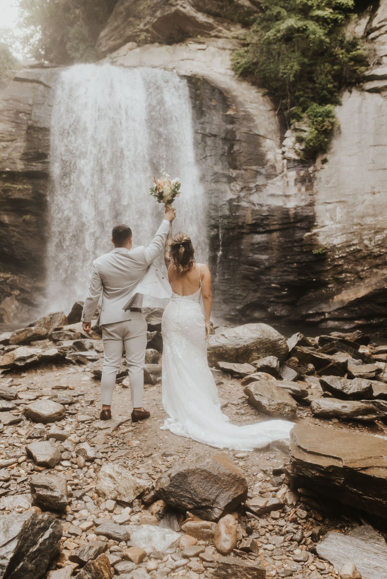 Man and woman hold hands and flowers in the air while facing waterfall during Asheville elopement.