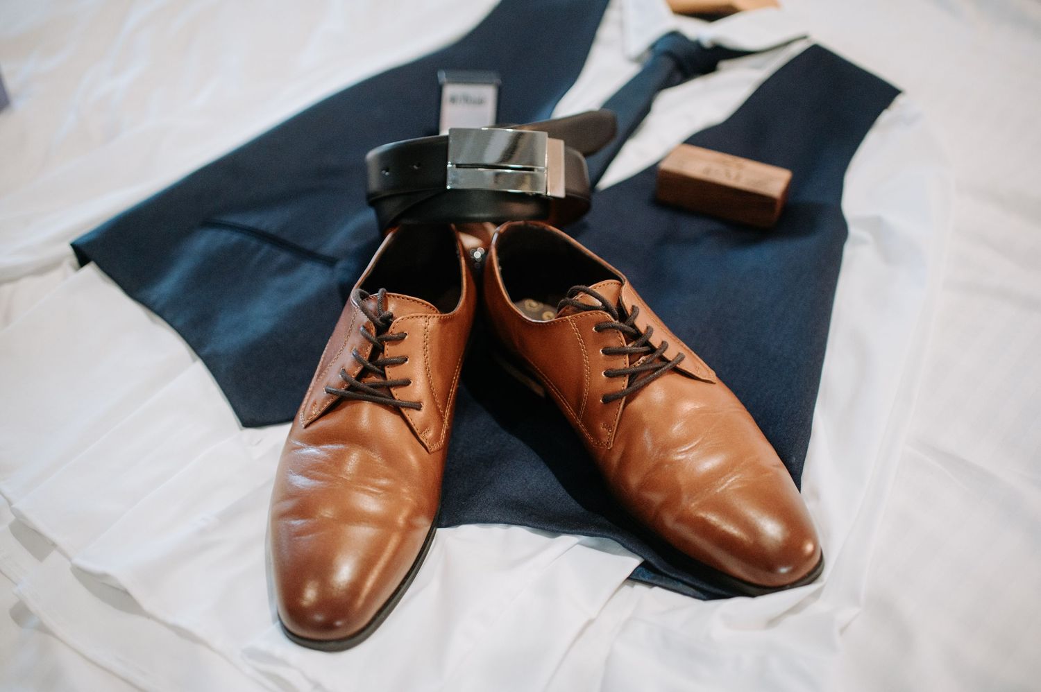 Brown leather dress shoes and navy vest laid out on white fabric.