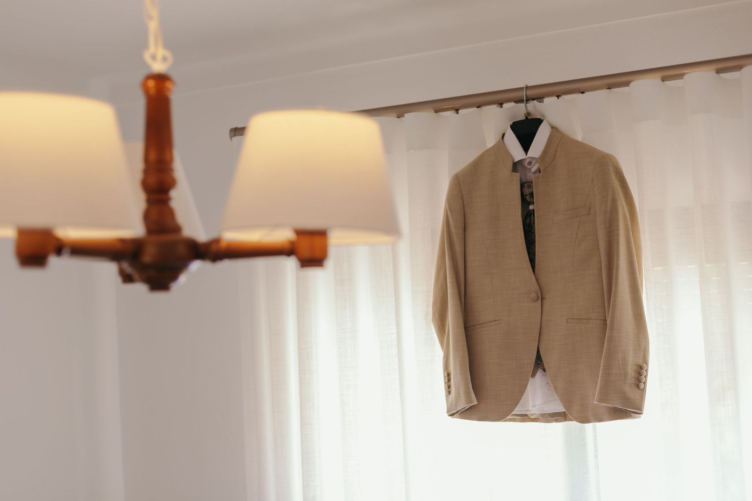 Beige wedding suit jacket hanging on curtain rod with elegant chandelier in background.