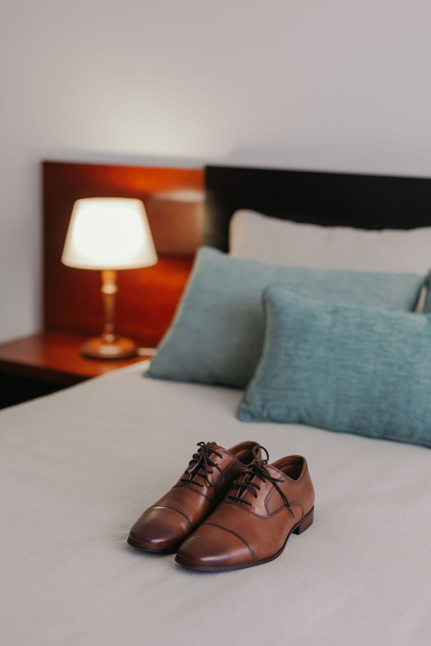 Brown leather Oxford dress shoes placed on a white hotel bed with teal pillows and glowing bedside lamp.