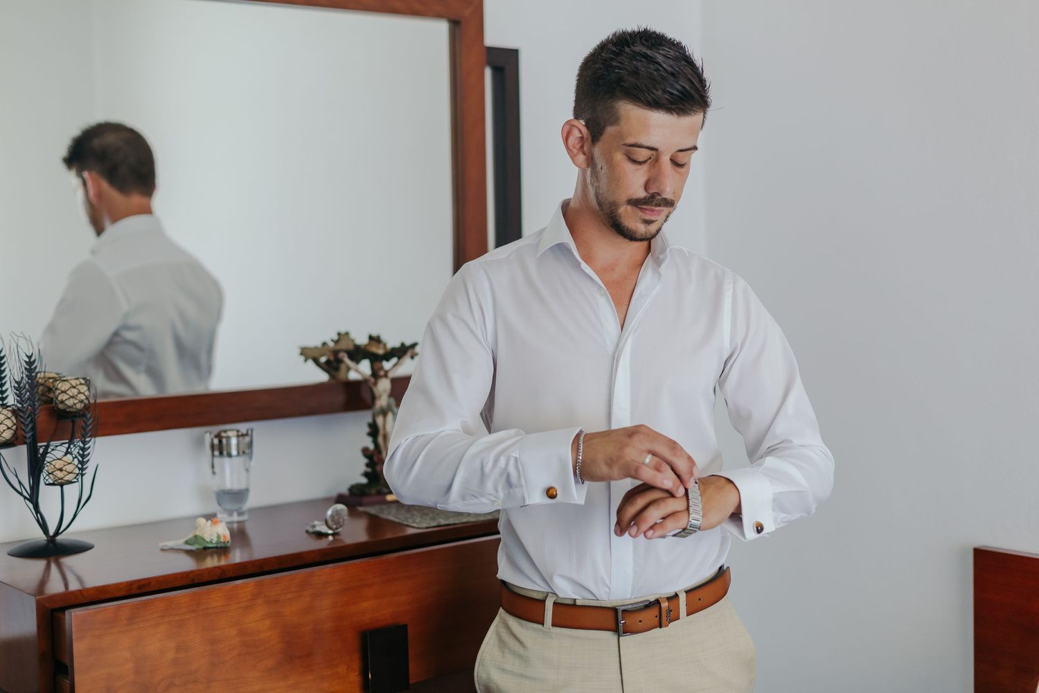 Groom in white shirt fastening cufflinks while standing before a wooden dresser mirror.