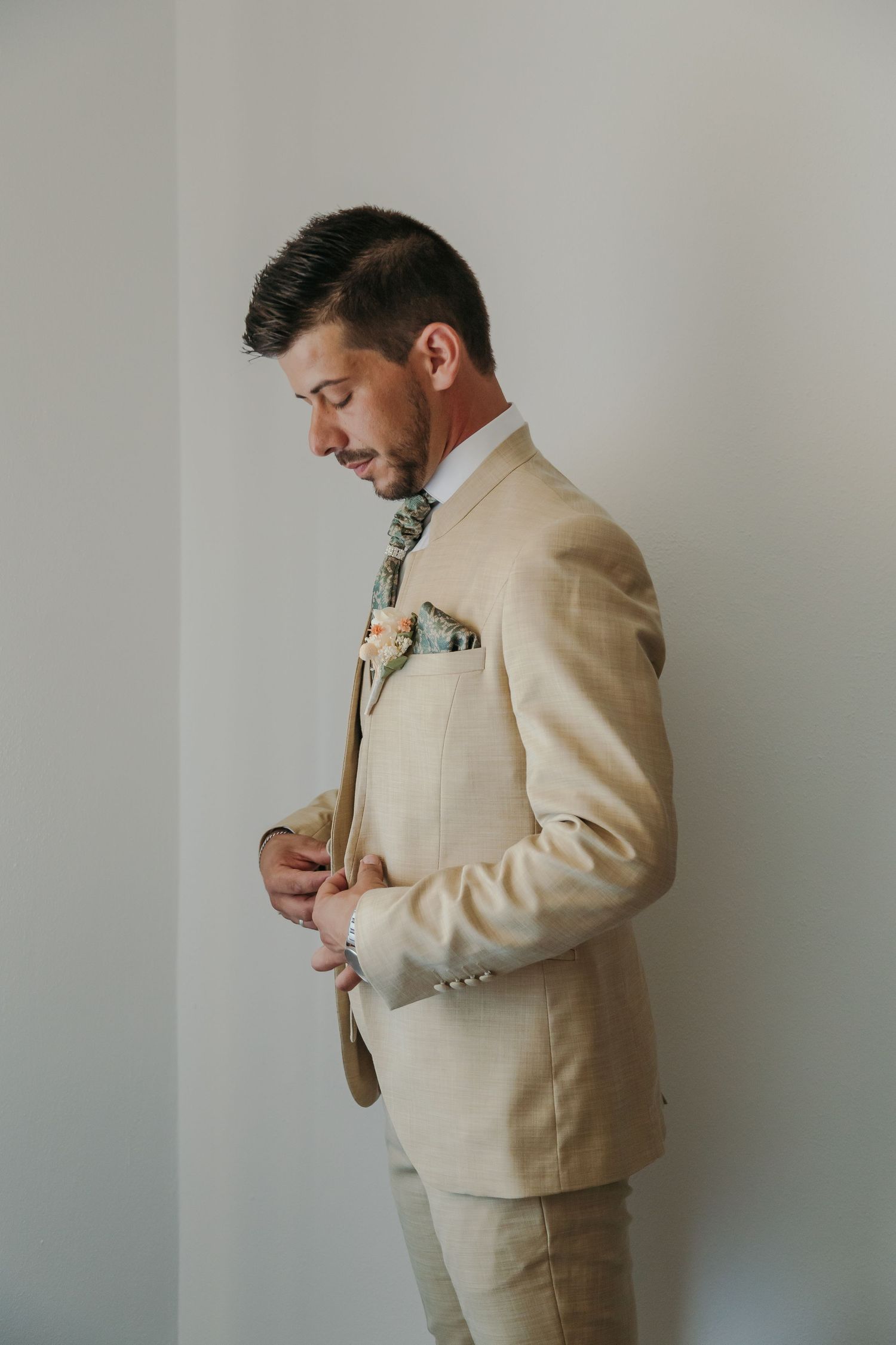 Groom in elegant beige linen suit adjusts cufflinks with floral boutonniere on lapel.