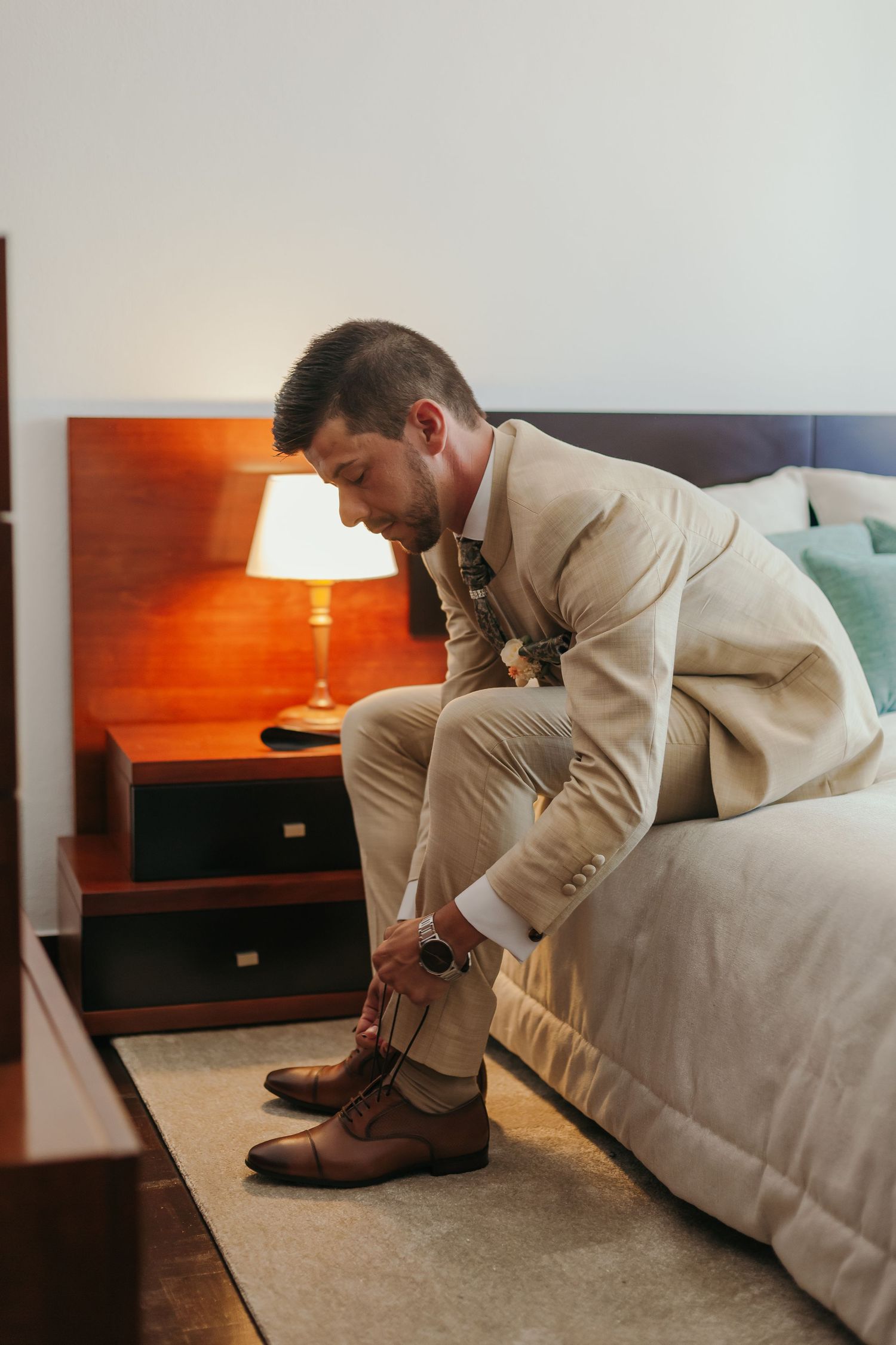 Groom in beige suit sits on bed lacing up brown leather dress shoes before wedding.