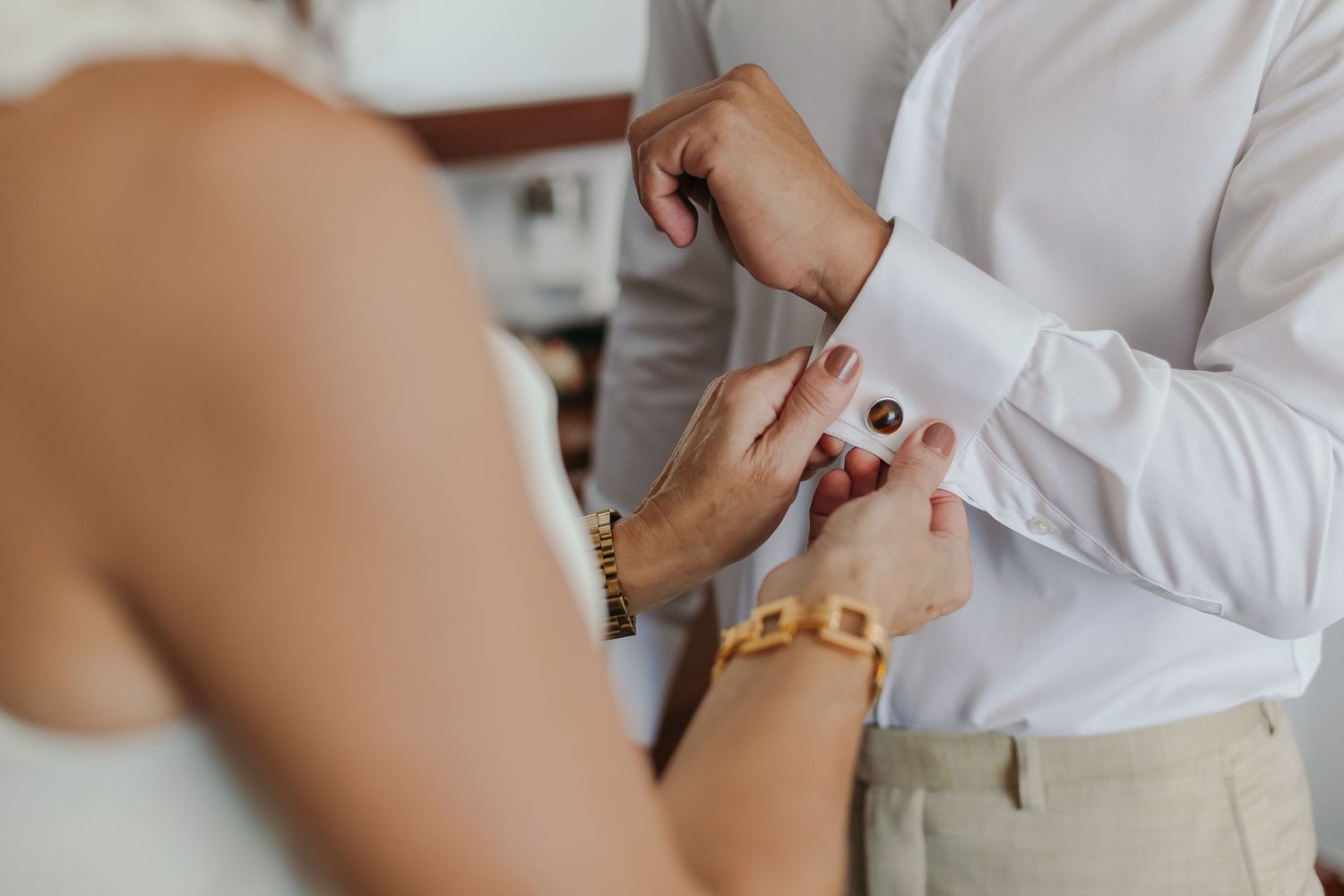 Bride helping groom fasten red cufflinks on white dress shirt before wedding ceremony.