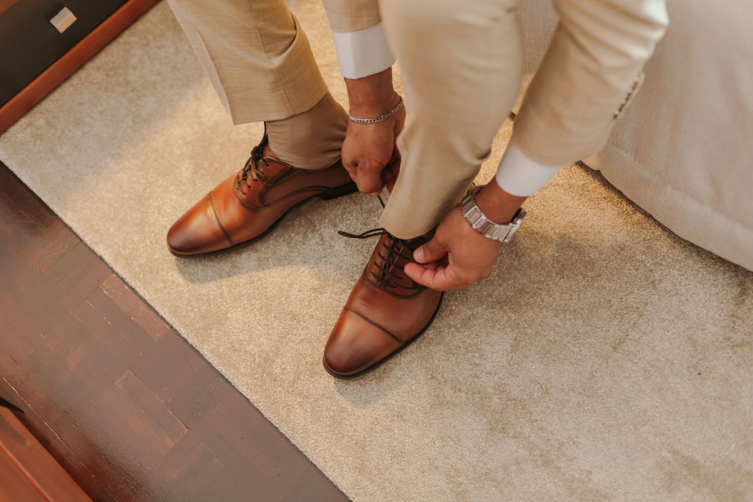 Groom in beige suit laces up brown cap-toe Oxford shoes on carpeted floor.