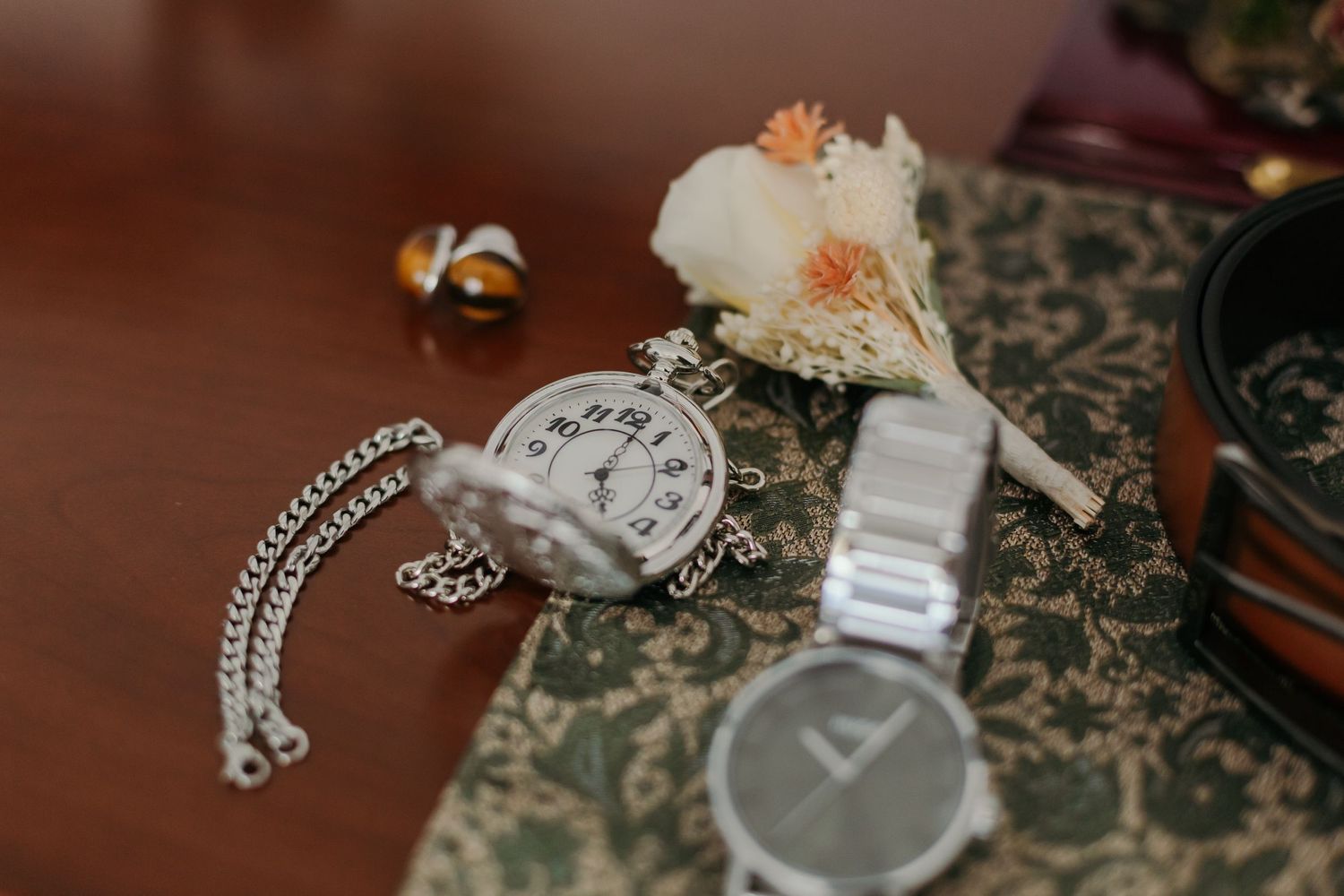Wedding flat lay featuring a vintage pocket watch, boutonniere, chain, and modern wristwatch on ornate fabric.