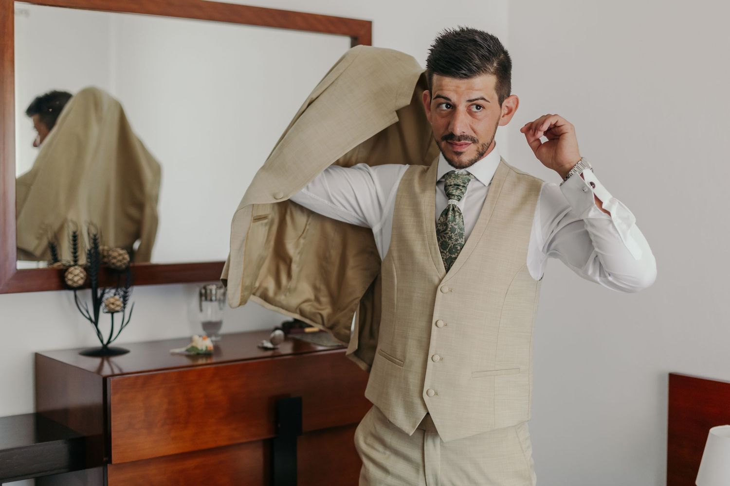 Groom in beige vest and white shirt puts on his suit jacket before wedding.
