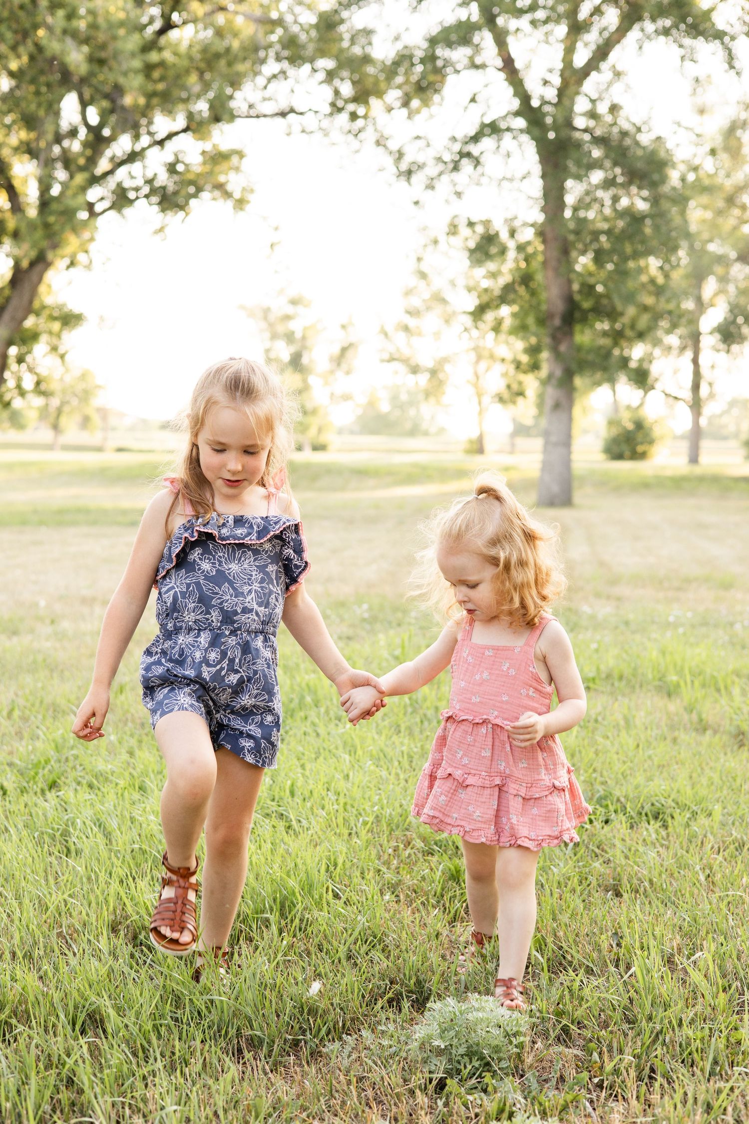 Two young children holding hands while playing in grassy field with trees.