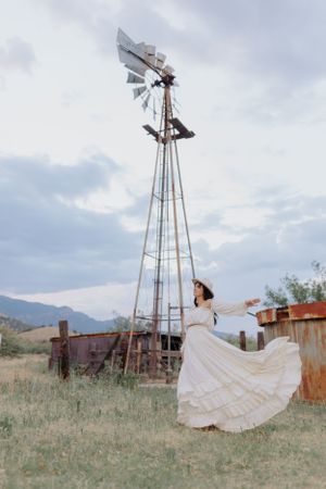White flowing dress billows in the wind against a rustic windmill and mountain landscape.