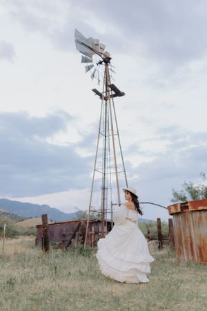 White dress twirls gracefully near an old windmill in a rural field setting.