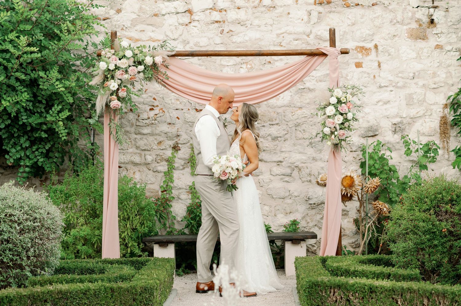 Couple shares their first look under a pink draped arch with stone wall and garden backdrop.