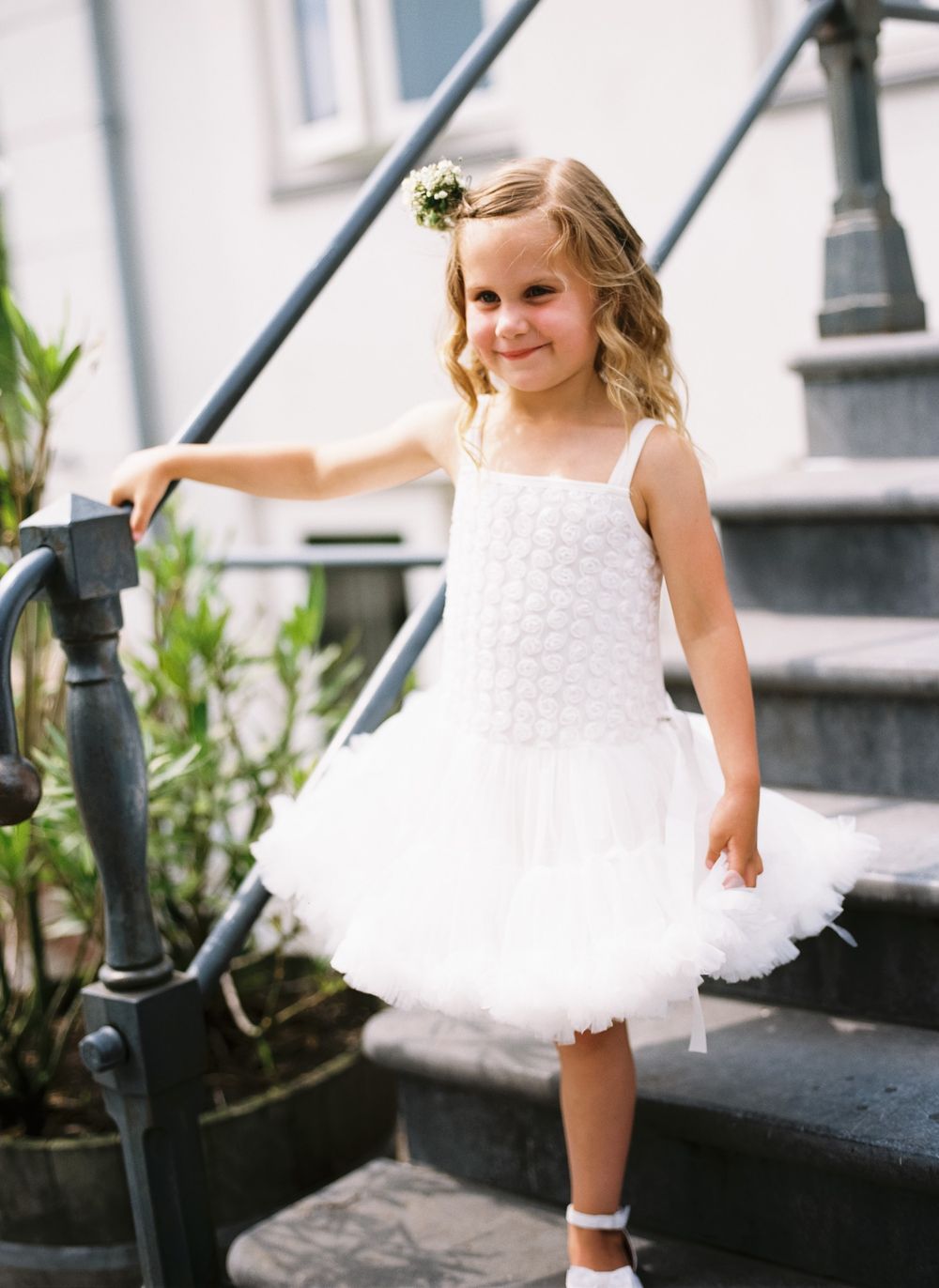 A young child in a white tulle dress stands on outdoor steps wearing a small floral hair accessory.