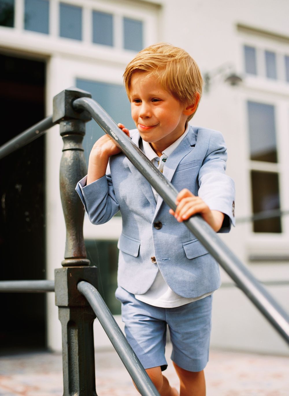 A child in a light blue blazer and shorts leans against an outdoor metal railing.