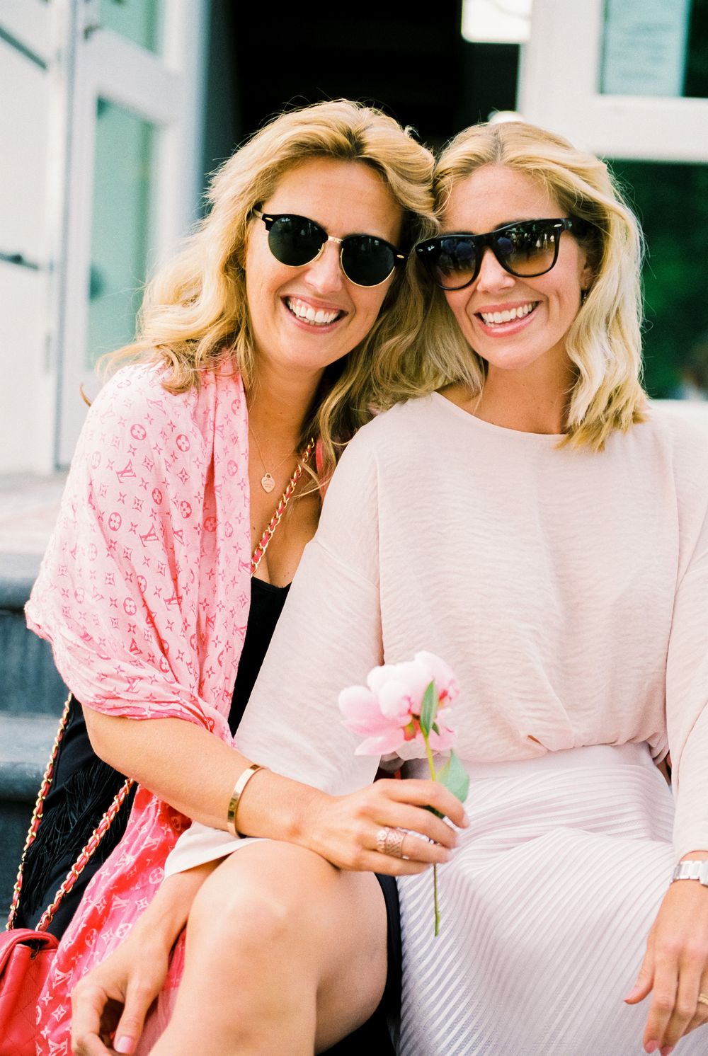 Two friends in sunglasses share a moment on steps while holding flowers.