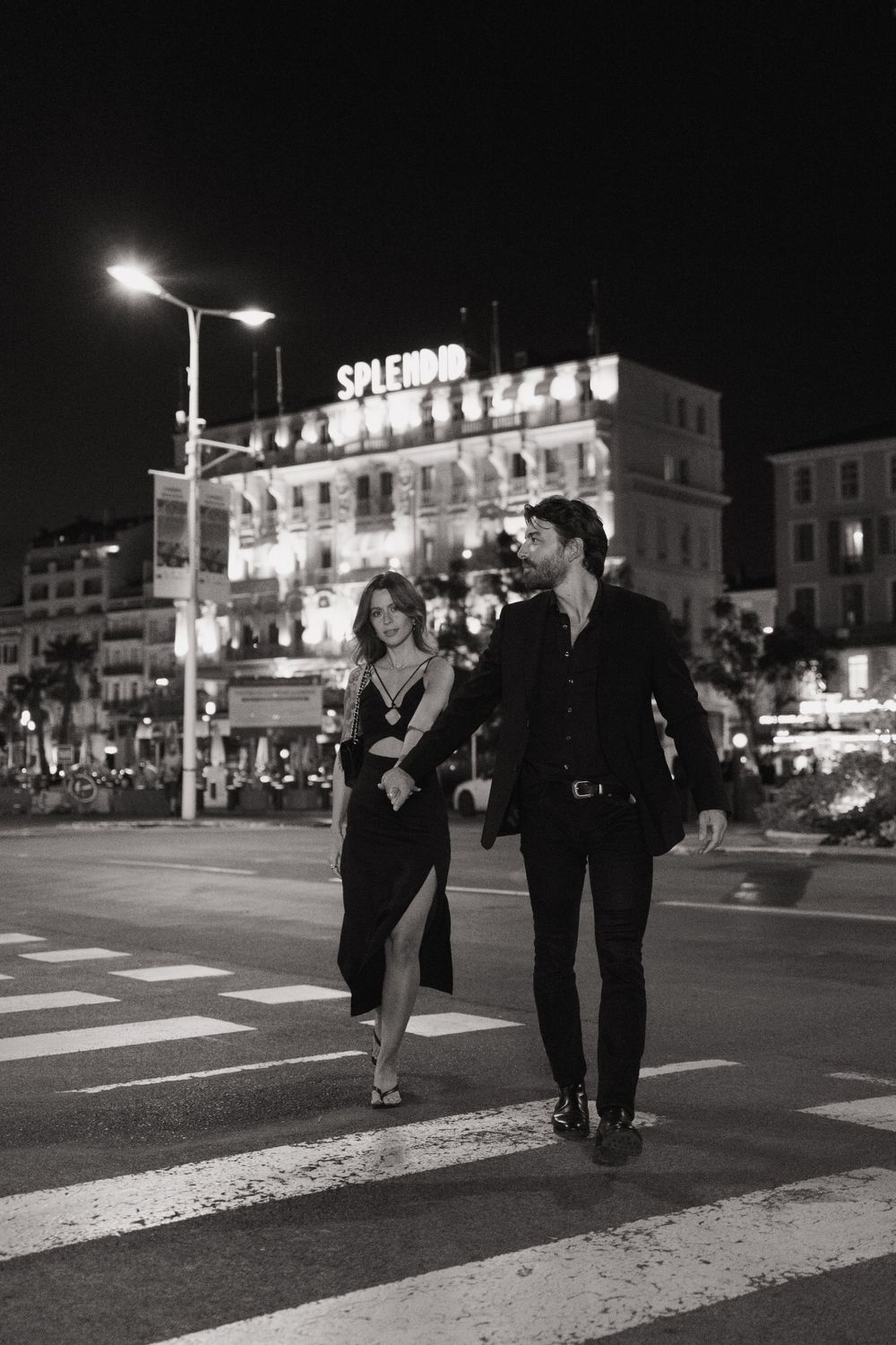 Couple walking across crosswalk at night with illuminated buildings in background.