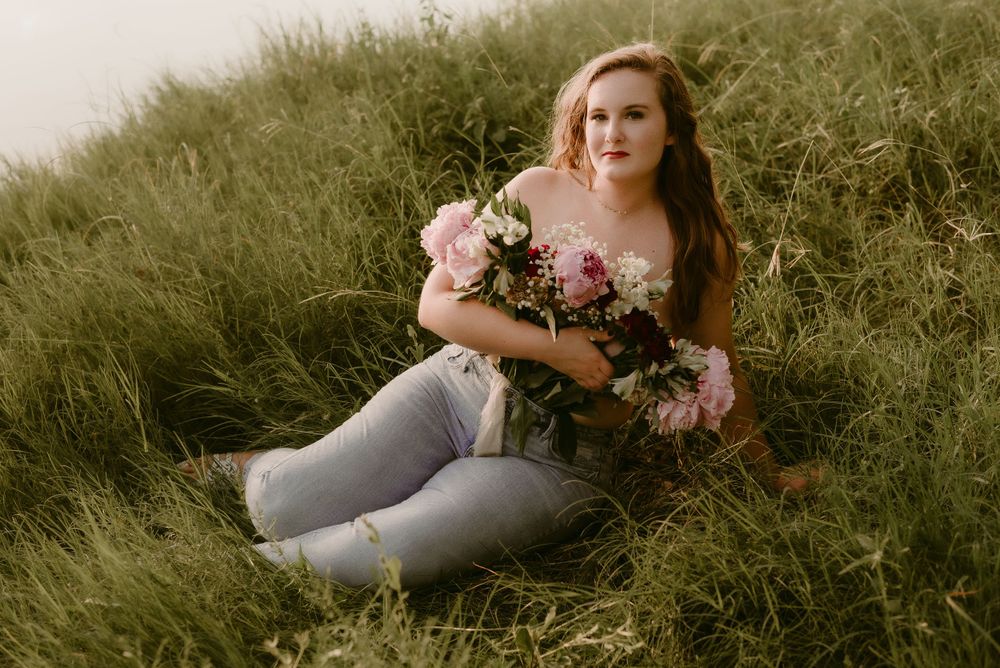 Relaxed outdoor portrait with someone sitting in tall grass holding a floral bouquet.
