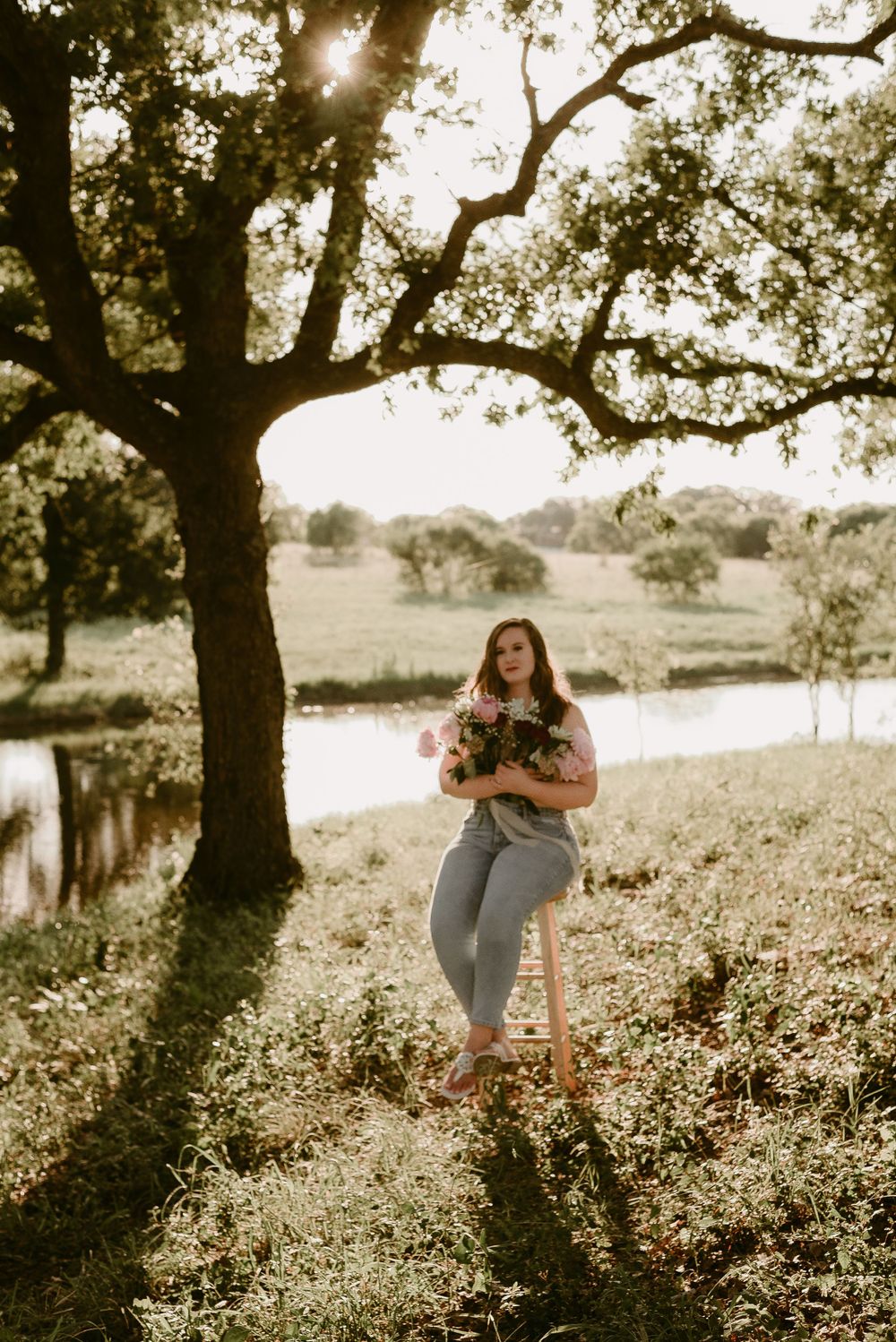 Sunlit nature portrait with someone holding flowers by a pond under a large oak tree.