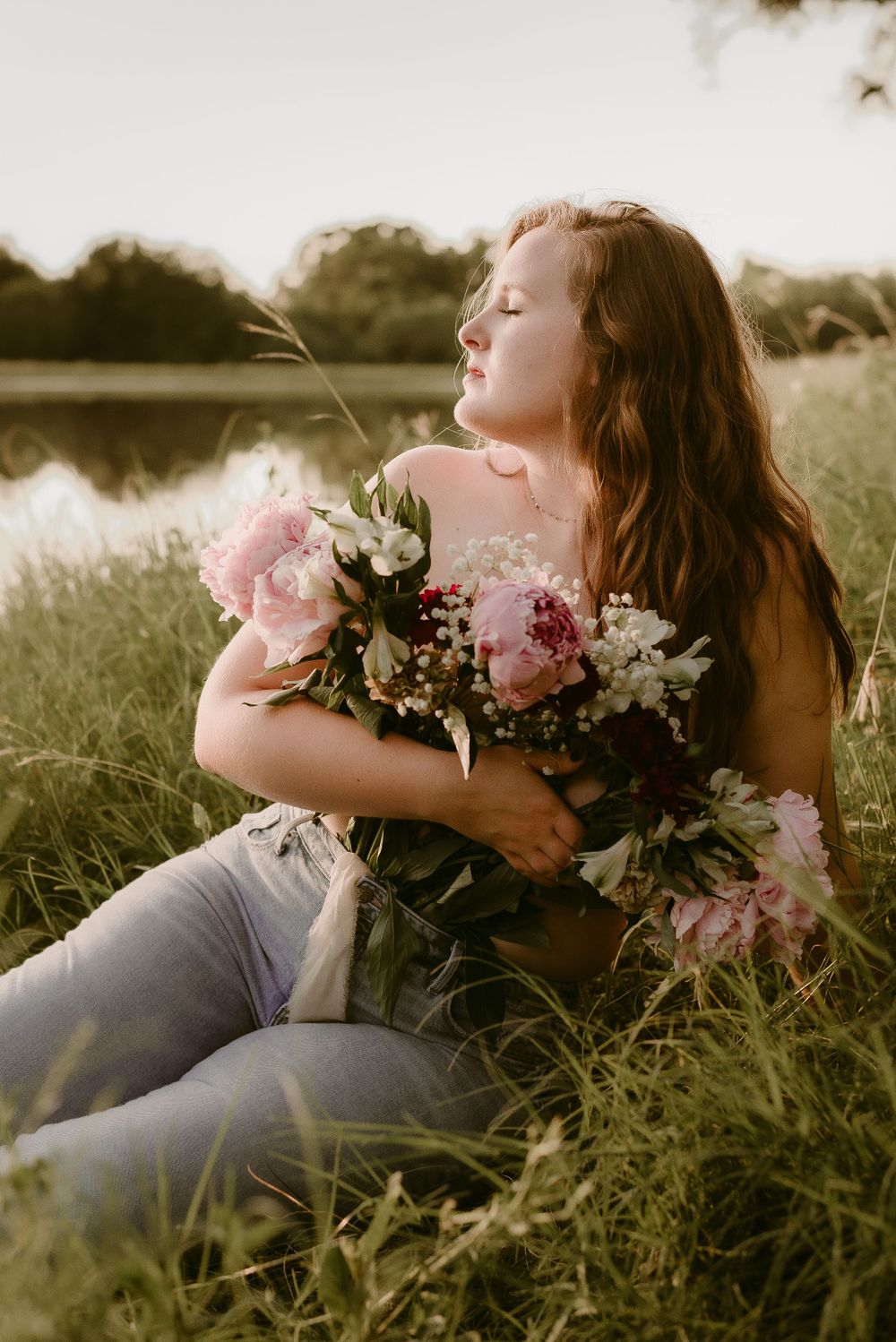 Romantic sunset portrait with someone embracing flowers while reclining in grass by a pond.