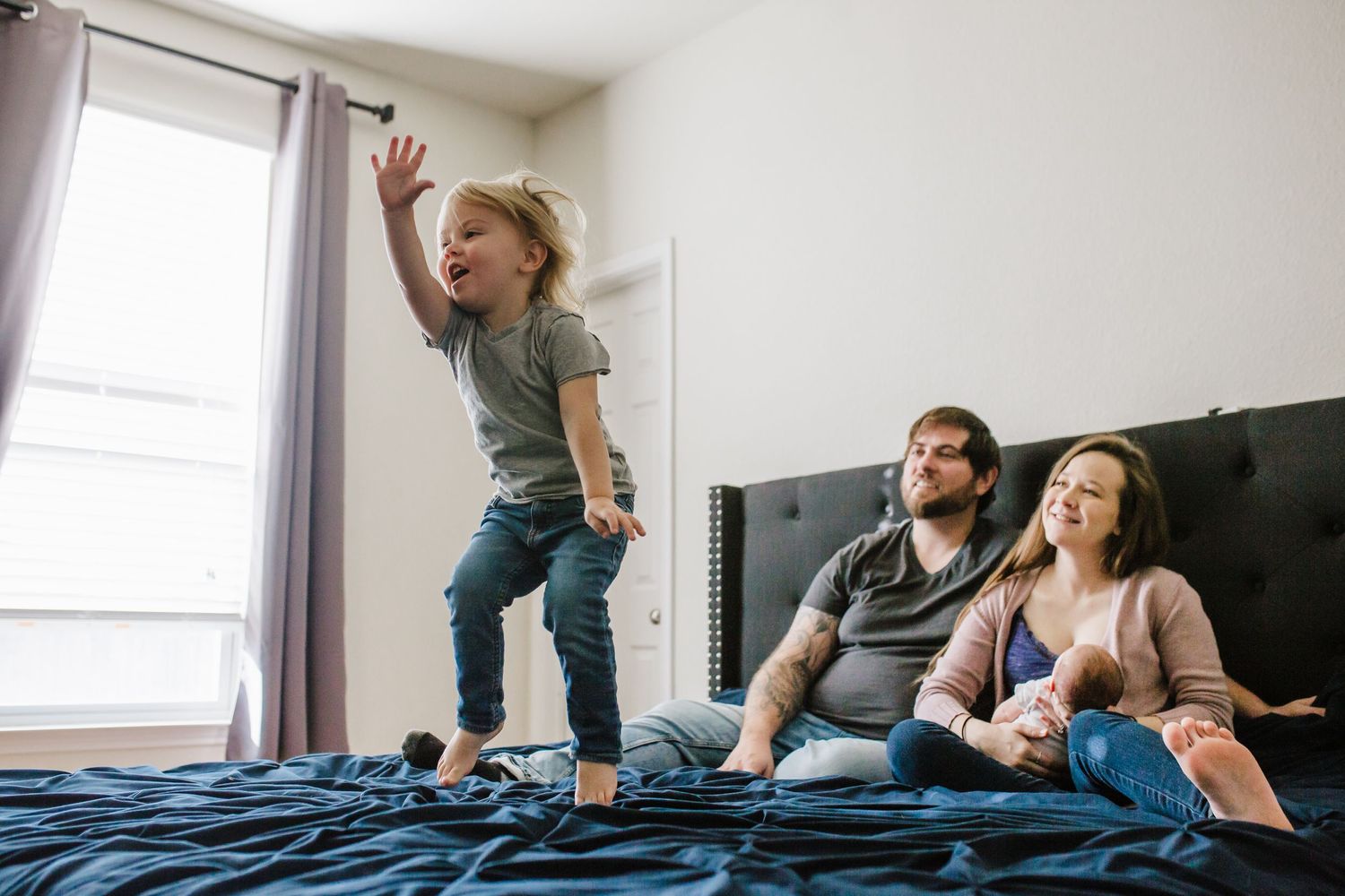 Young boy jumps on the bed while his parents hold newborn baby and look on