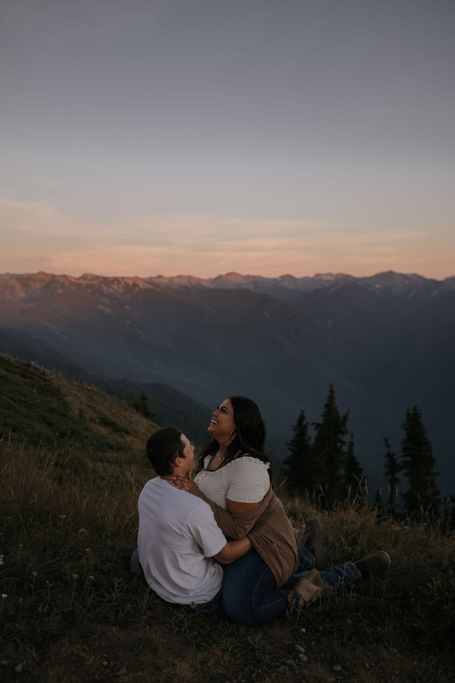 A couple sits on a hillside overlooking mountain ranges at sunset.