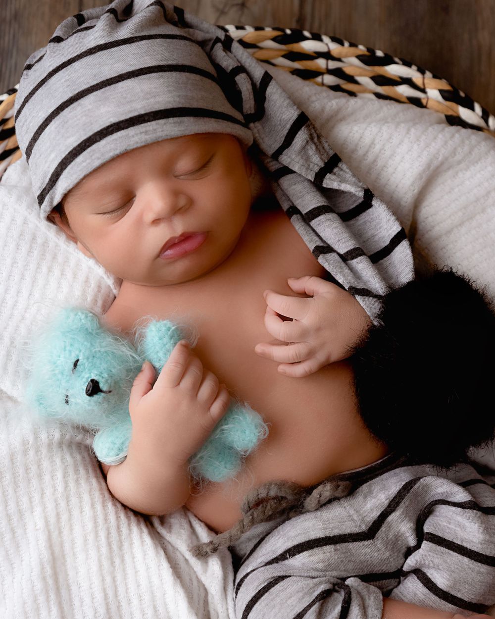 Sleeping baby in striped cap holding small blue teddy bear on white blanket.