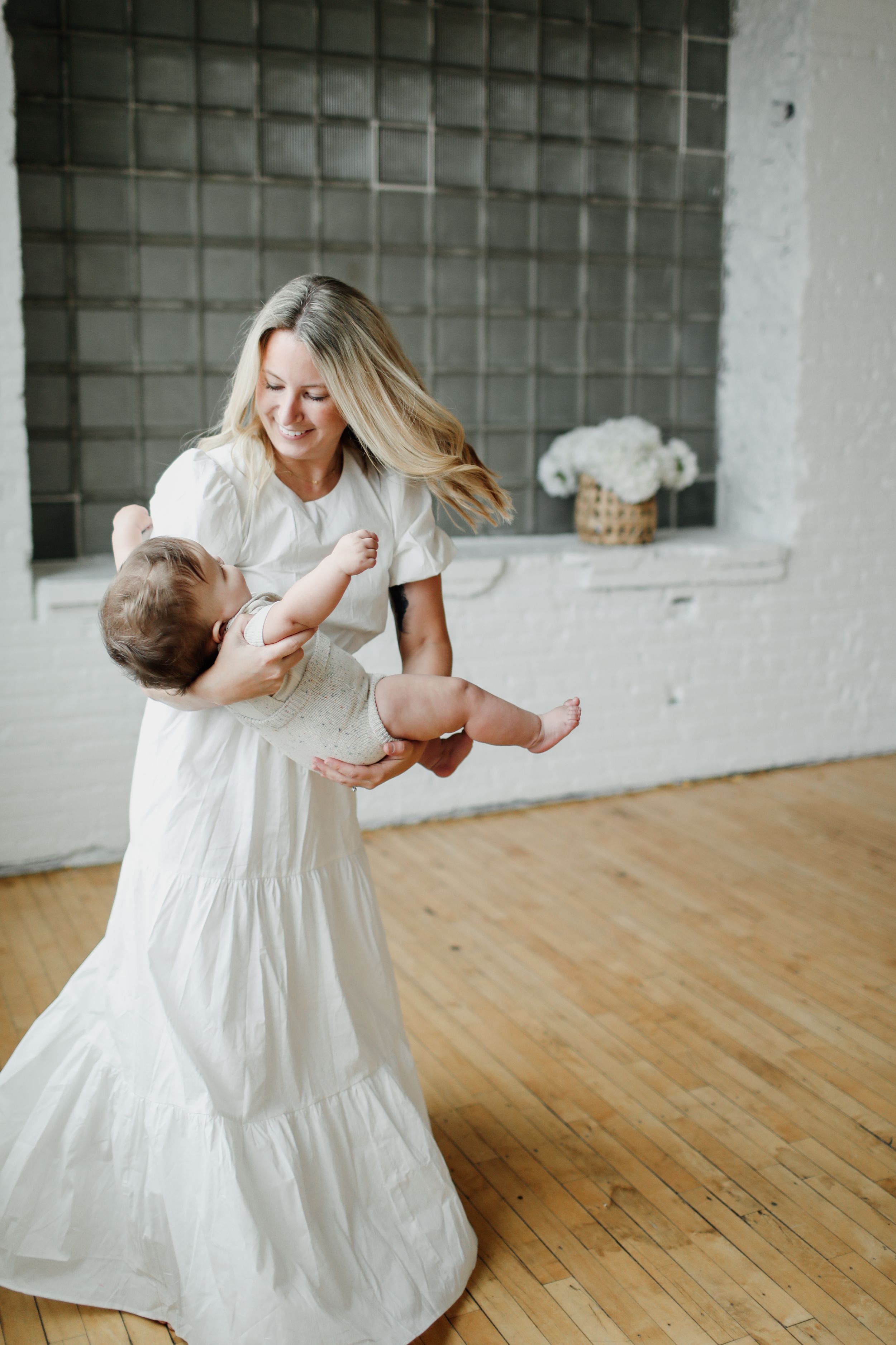 Person in white dress dancing with baby in bright studio space.