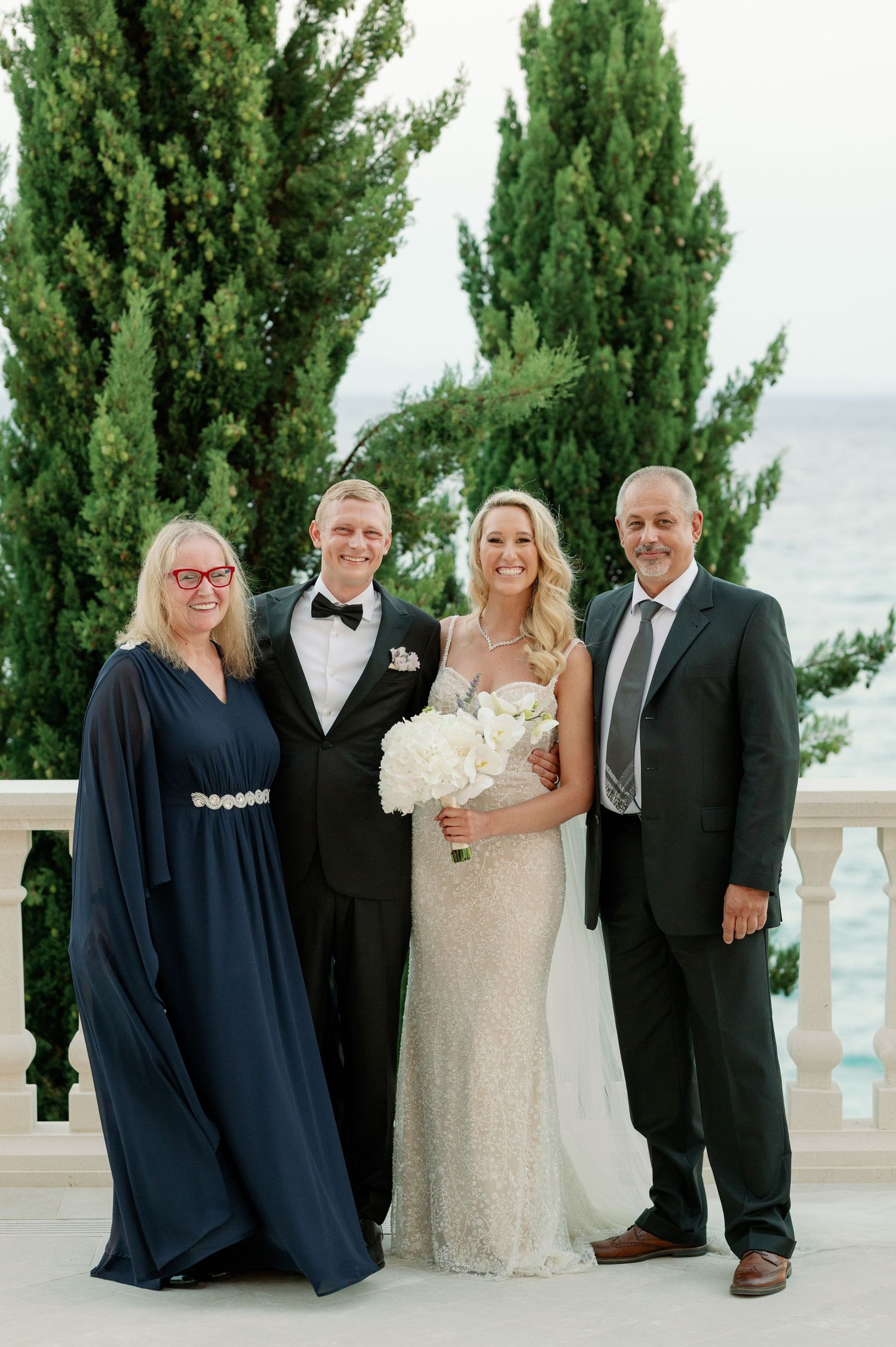 Wedding group photo on elegant balcony with cypress trees and ocean view in background.