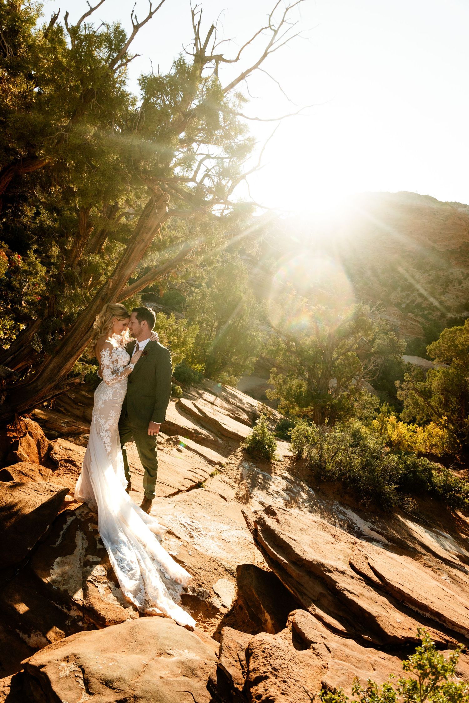 Wedding couple embracing on red rock formation with sunburst through trees.