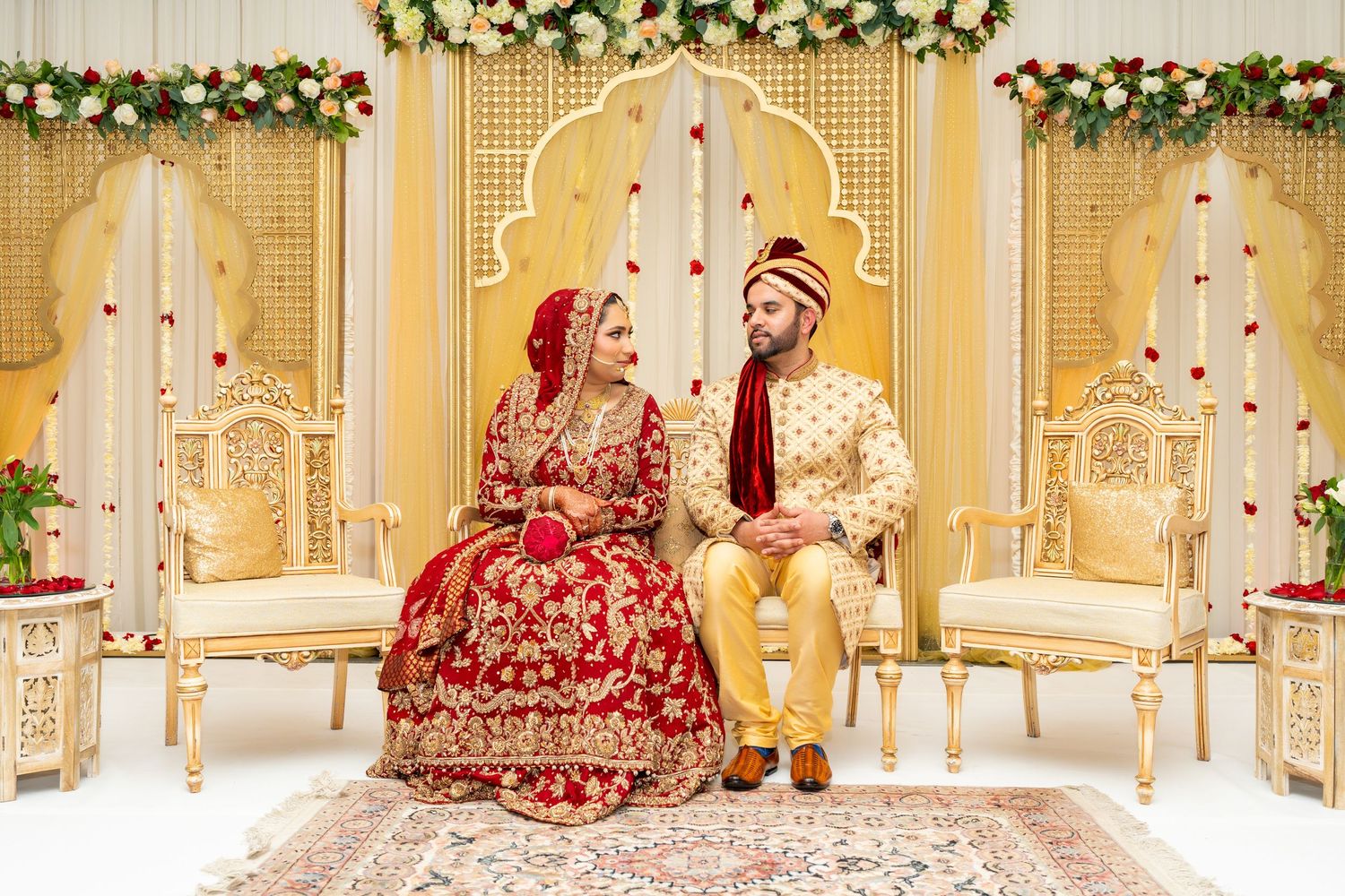 Newlywed couple in traditional red and gold attire seated on ornate chairs at wedding venue.