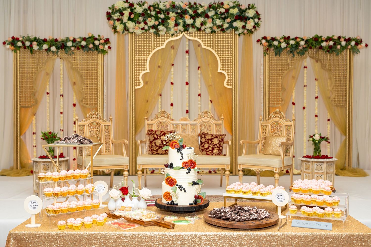 Elegant wedding dessert table with tiered cake and treats against gold and white backdrop.
