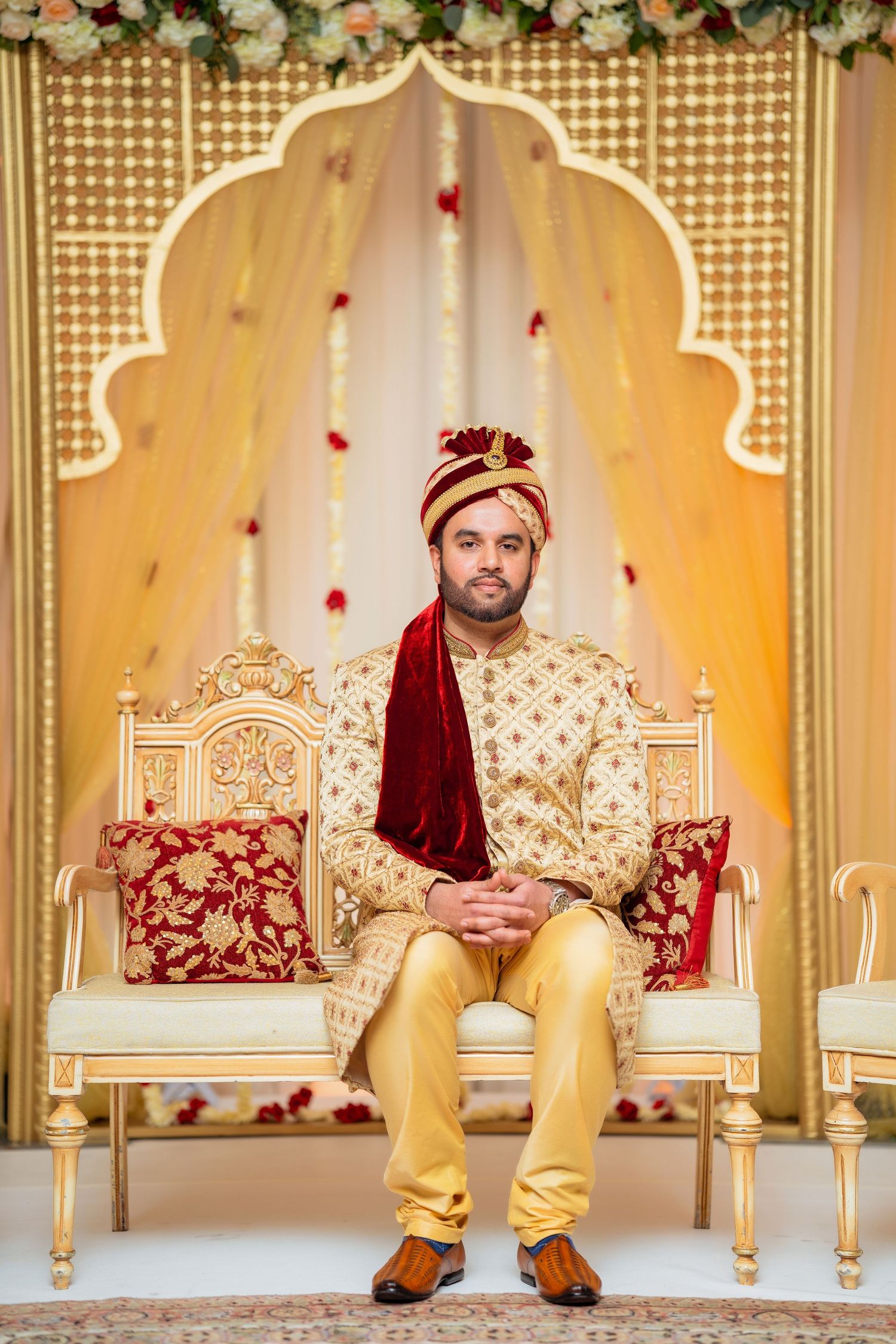 Elegant Indian wedding setup with ornate throne chair against gold archway decorated with rose petals.