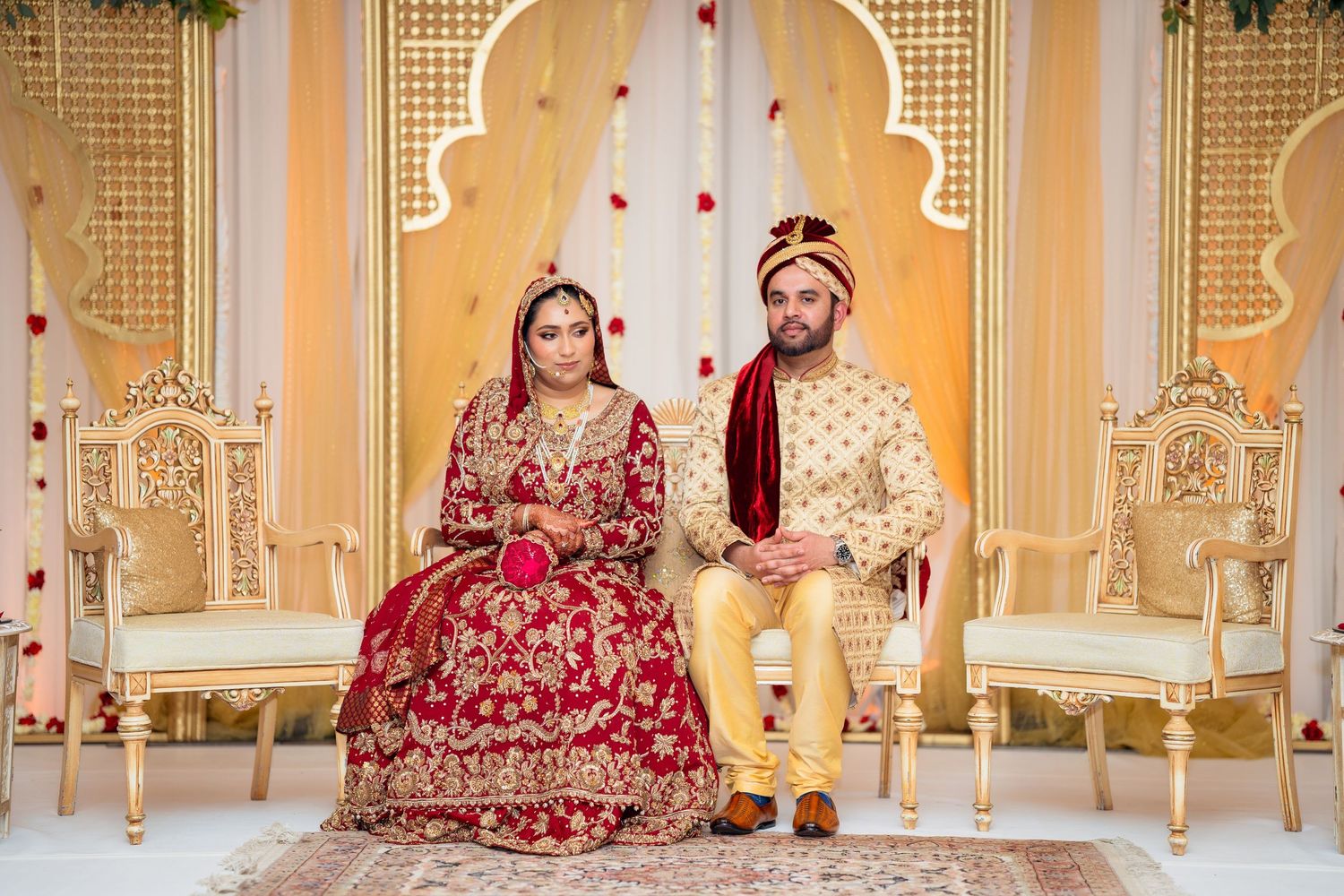 Traditional Indian wedding couple seated on decorative gold thrones against ornate backdrop.