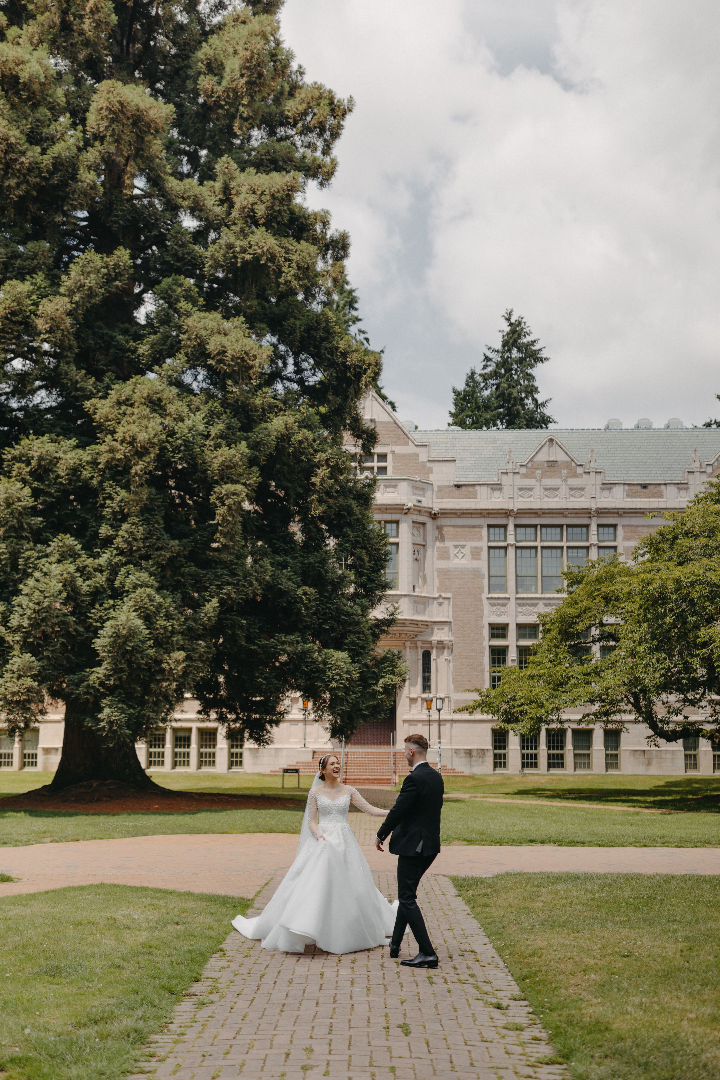Wedding couple poses in front of historic university building surrounded by majestic trees.