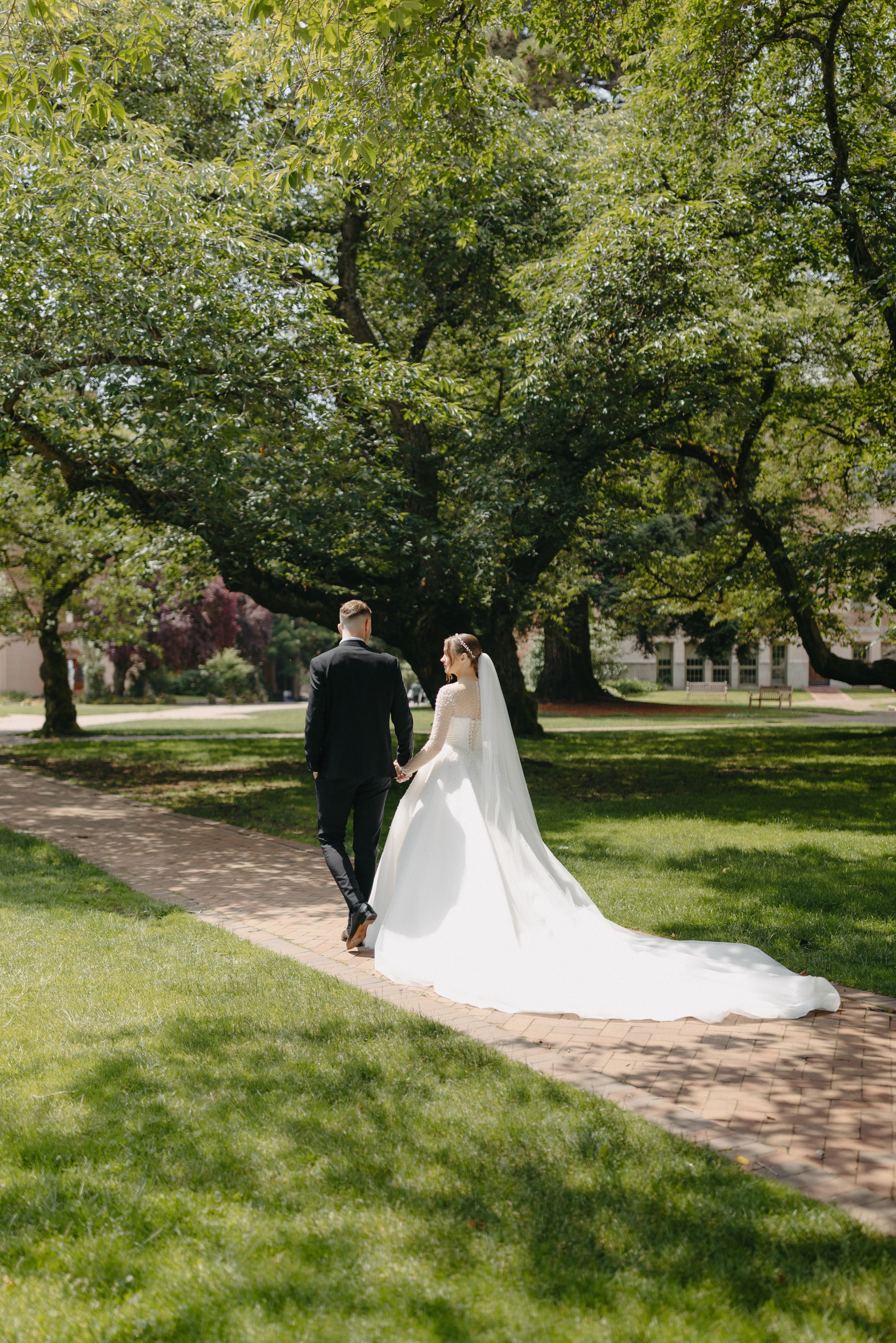 Newlyweds walking down tree-lined path with bride's long train flowing behind them.