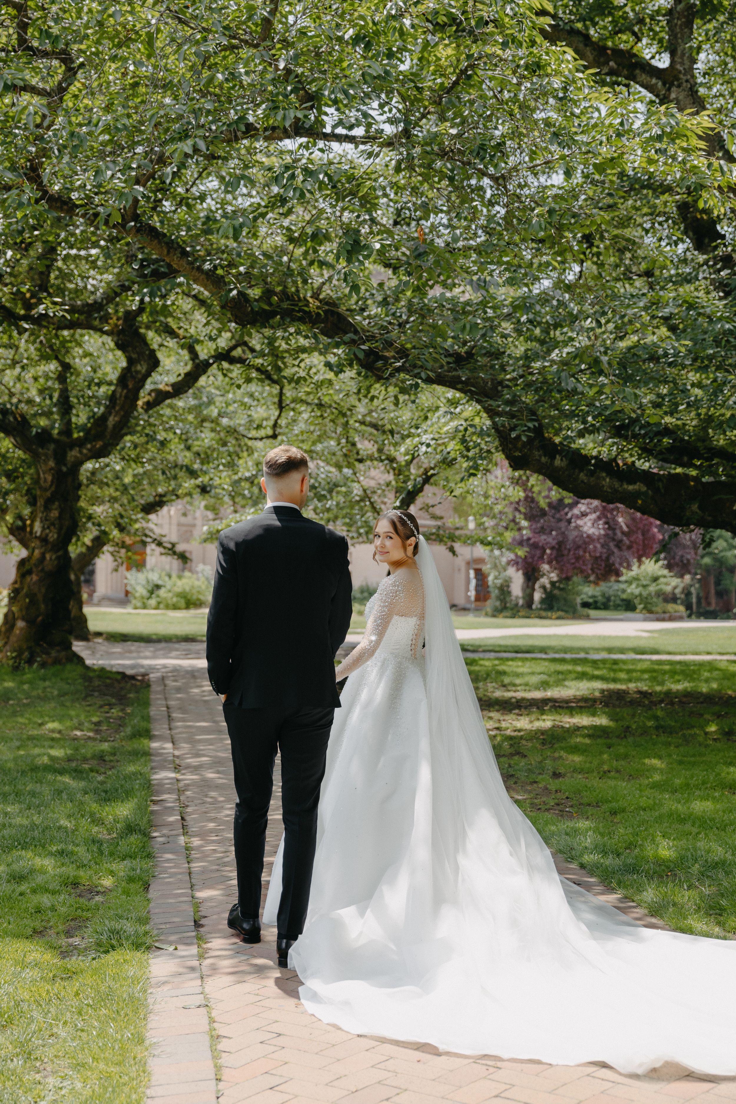 Wedding couple walking down brick path under canopy of green trees.