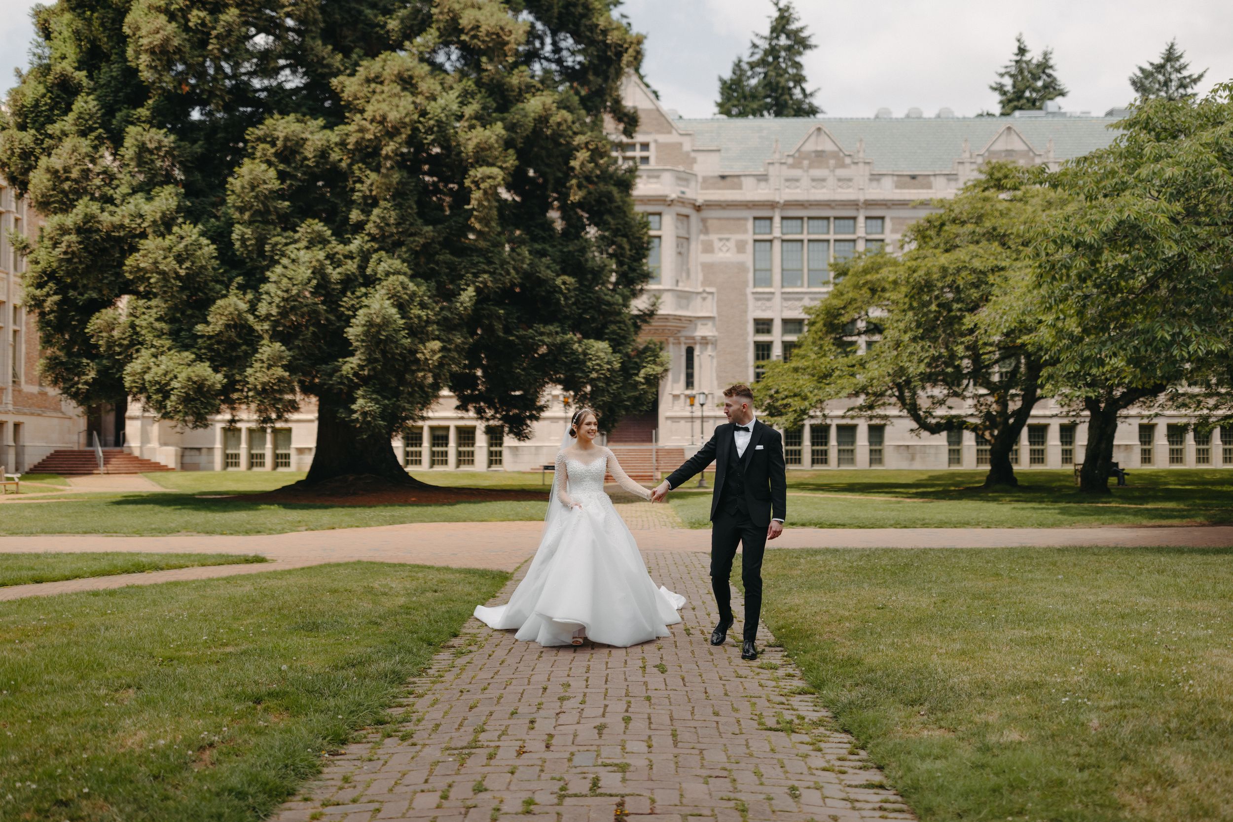 Bride and groom walk hand in hand across sunny campus lawn with classical architecture backdrop.