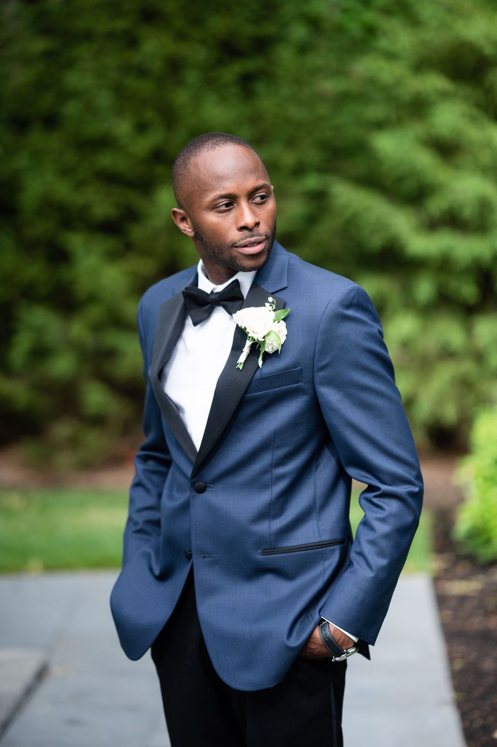 Stylish groom in navy tuxedo and bowtie poses outdoors against green foliage.